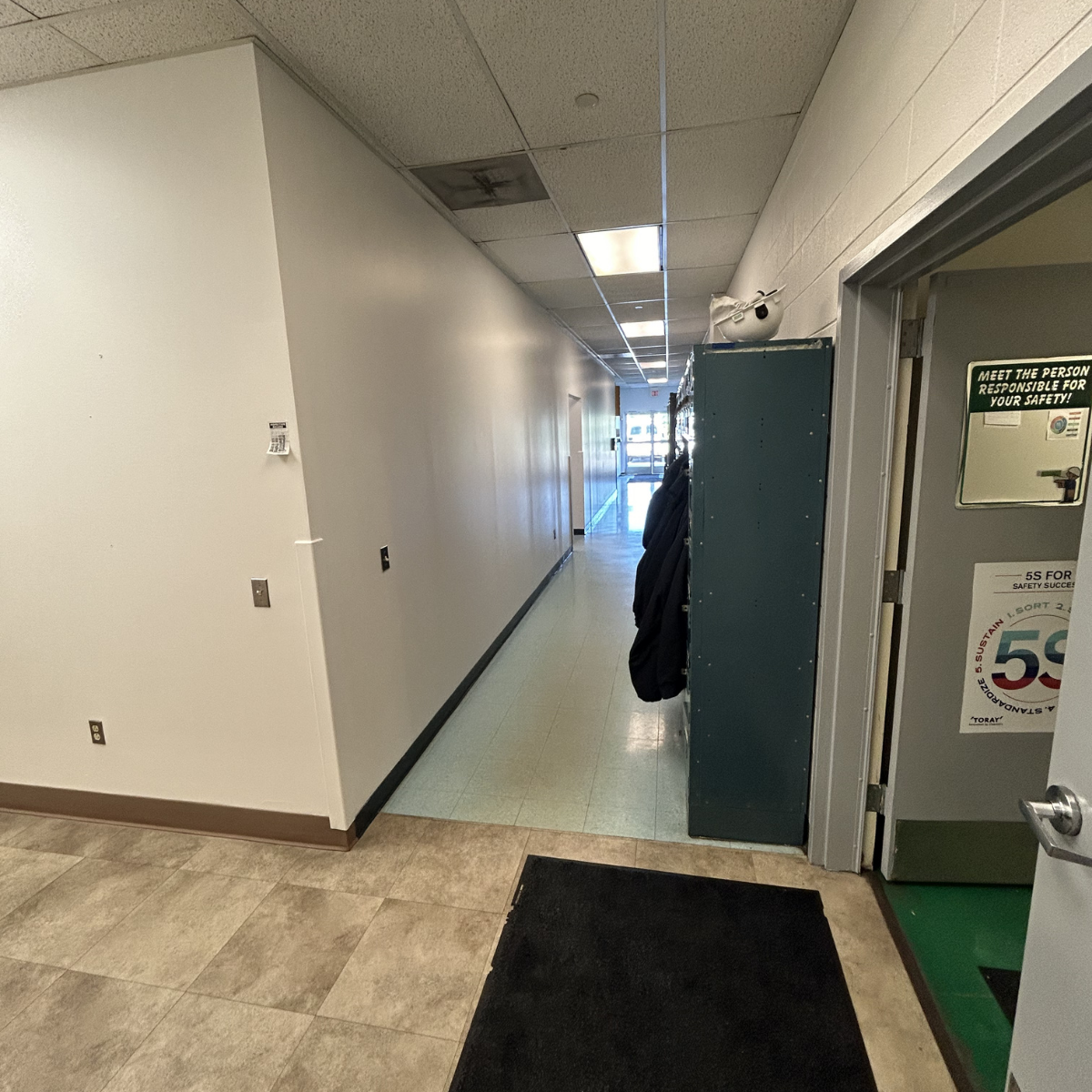 Winchester commercial painters CMT Painting Co. An indoor hallway leading to a glass door entrance, with a row of lockers on the right and a door with a safety sign, with beige tiled floor and white walls.