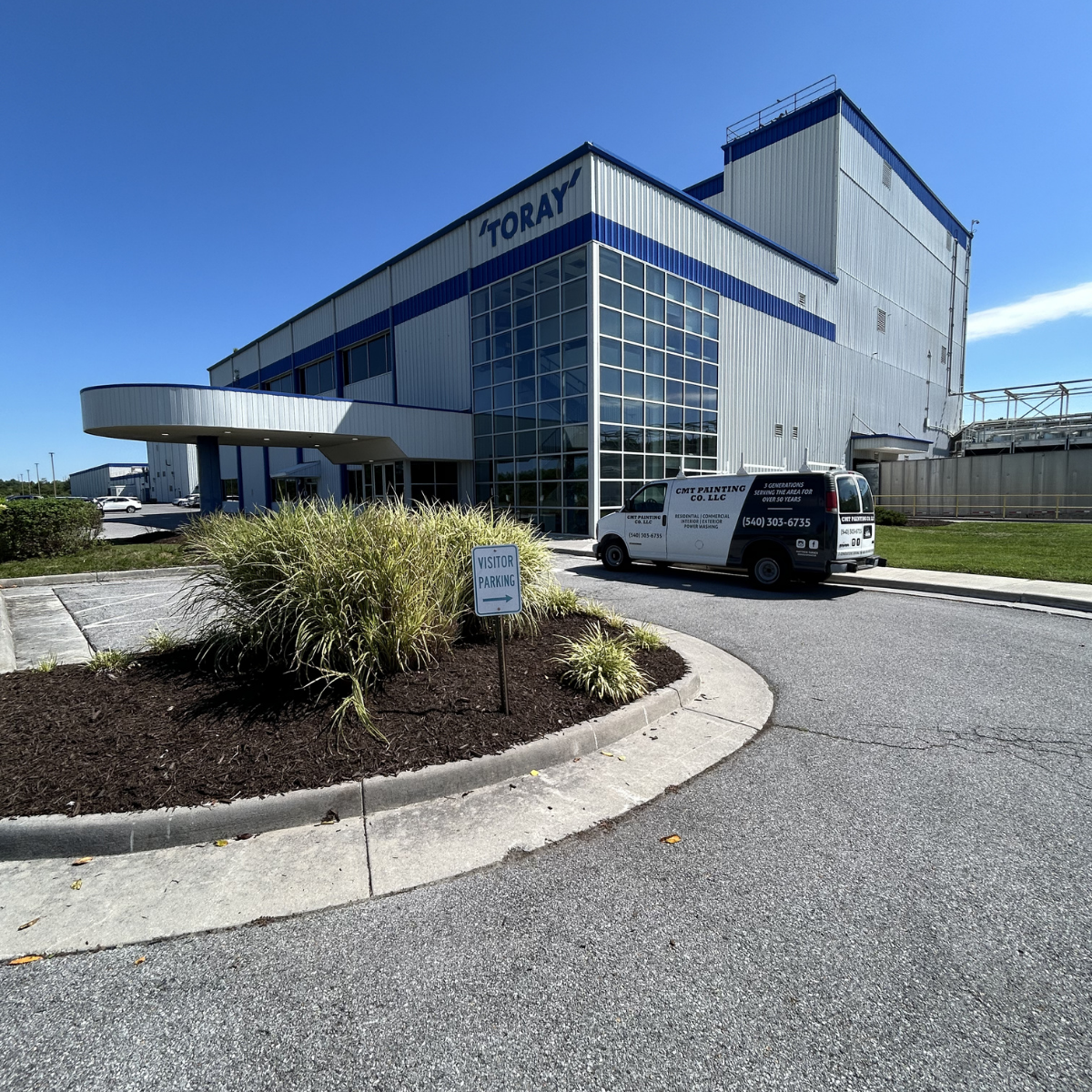 An industrial building with a sign reading 'TORAY' on the upper part of the structure. A van with company information parked in front, and a designated visitor parking area sign nearby. The building features large glass windows and a modern design, w