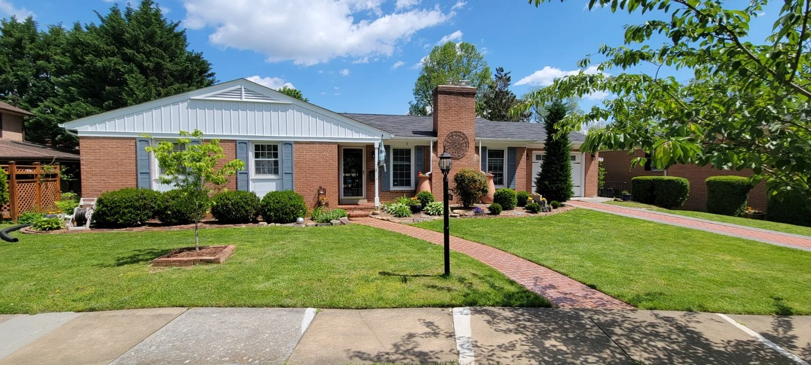 CMT Painting Co Exterior Painting Project in Winchester, VA. Front view of a brick house with a well-maintained lawn, brick walkway, and various bushes and trees under a partly cloudy sky.