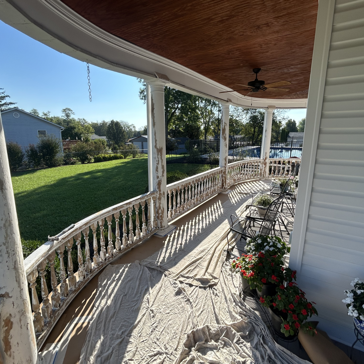 Front porch with potted flowers, white painted railing, ceiling fan, and a view of yard with grass and trees in the background.