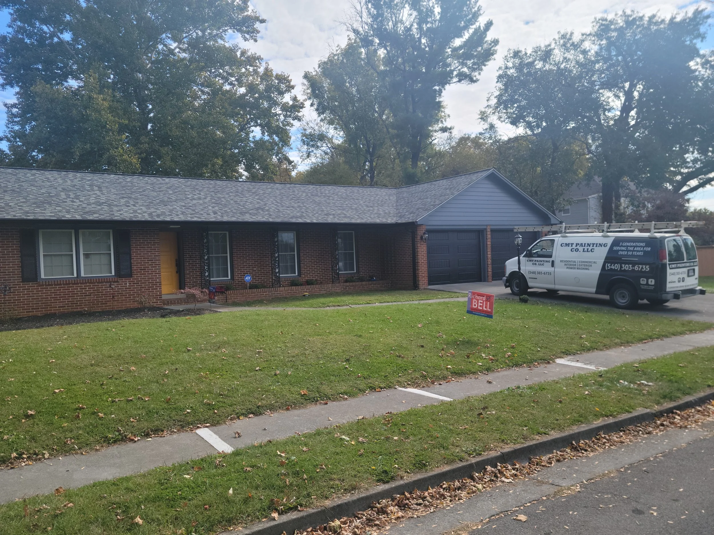 A single-story brick house with gray siding on the garage. There are four windows and a yellow front door. A white van with business branding is parked in the driveway, and there is a red political yard sign in the lawn. The yard has green grass and 