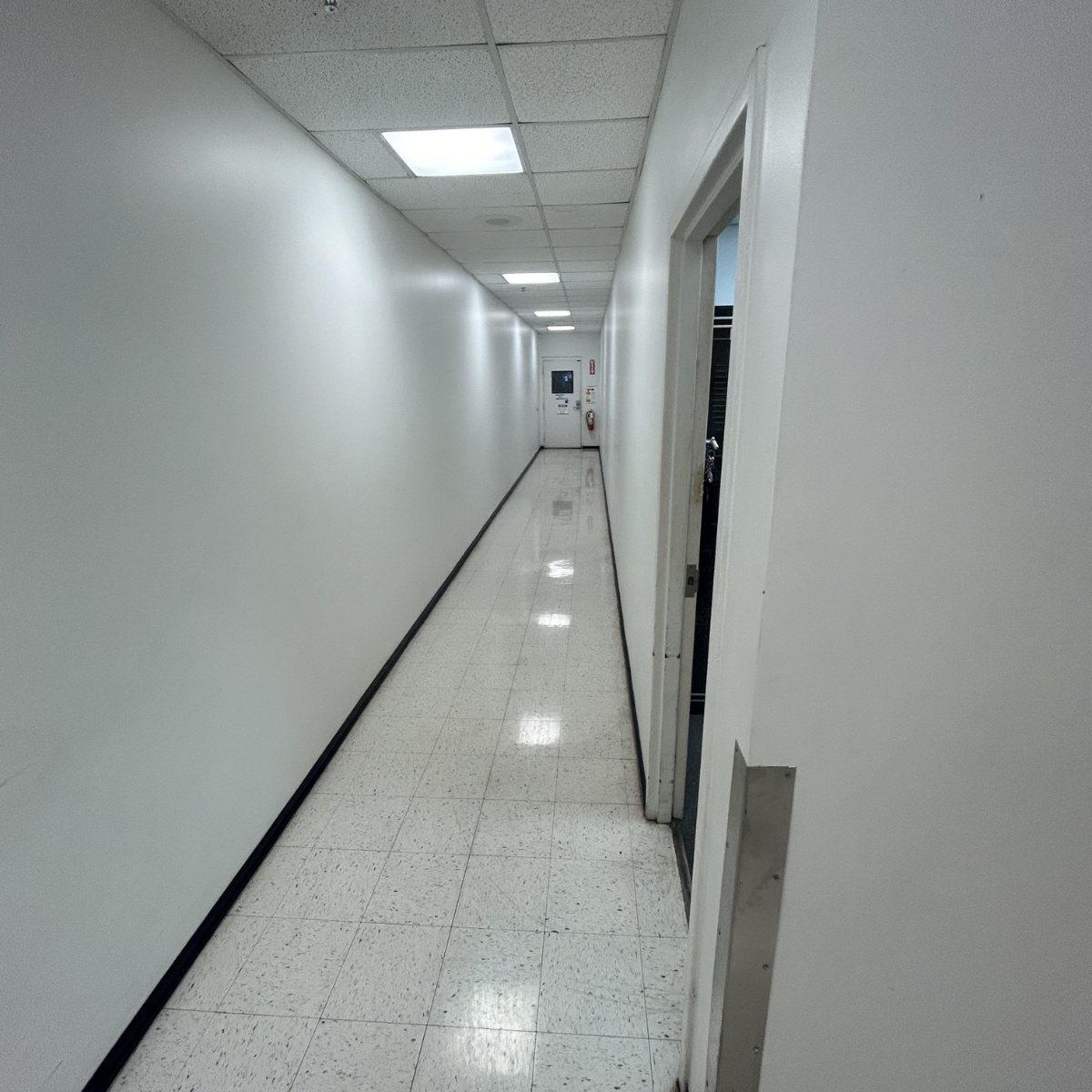 A long, narrow hallway with white walls, tiled flooring, and ceiling lights, leading to a door at the end.