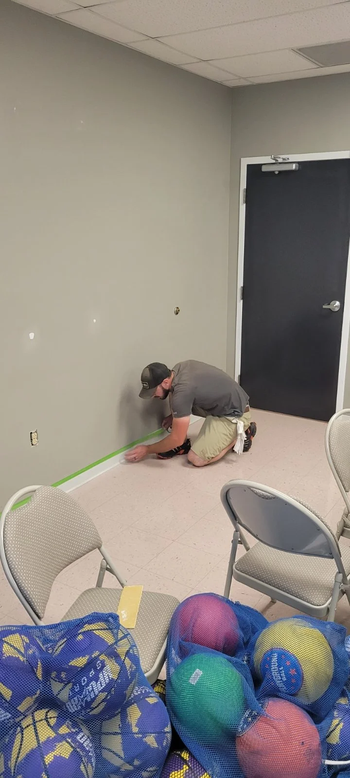 A man kneeling on the floor taping a green painter's tape along the baseboard of a gray wall in a room that appears to be under renovation. There are empty chairs in the room and a bag of sports balls in the foreground. CMT Painting Winchester