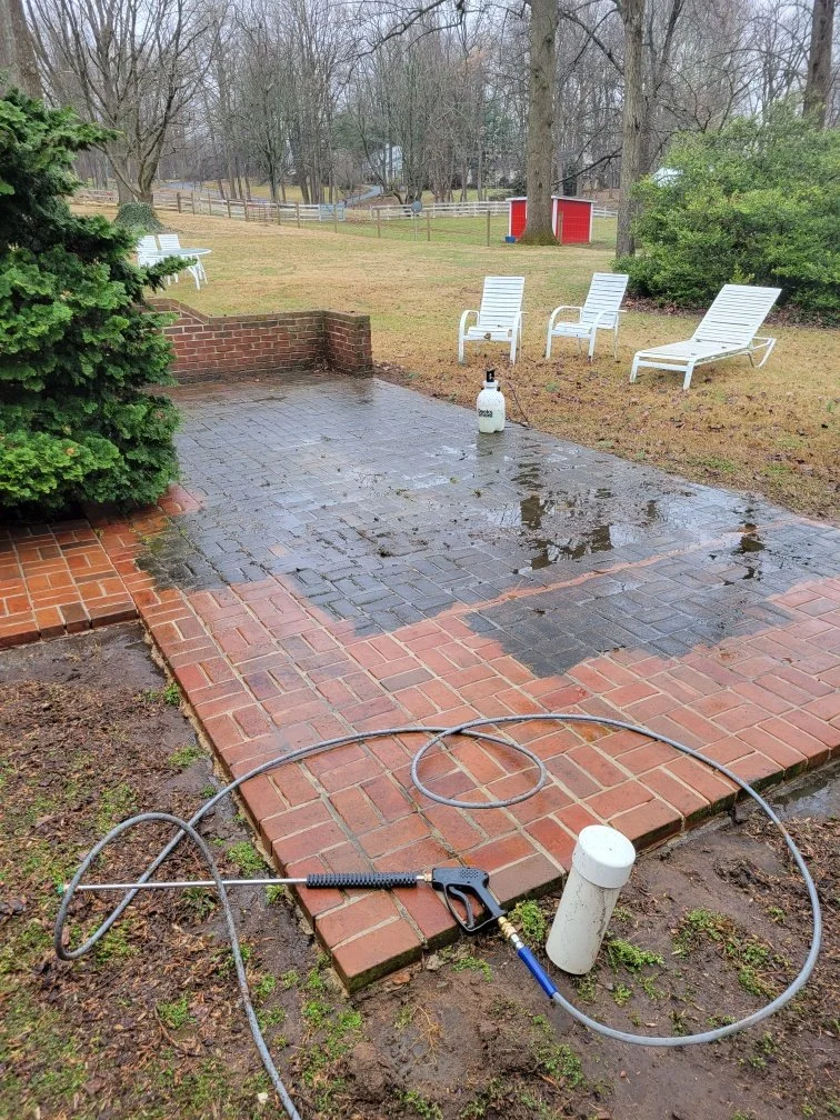Pressure washing Winchester Wet brick patio with a pressure washer hose and spray gun on the ground. Several white outdoor lounge chairs are on the grassy yard, with some water puddles on the patio. A red shed and trees are visible in the background.