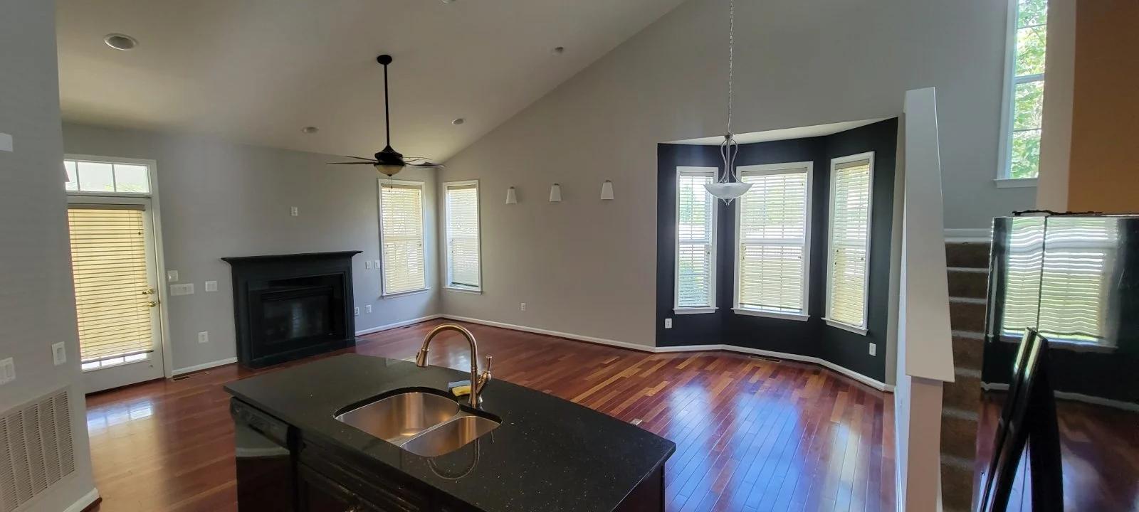 Winchester VA interior painting CMT Painting Co. Empty living room with hardwood floors, multiple large windows with blinds, a black fireplace, a ceiling fan, and a dark accent wall near a staircase.