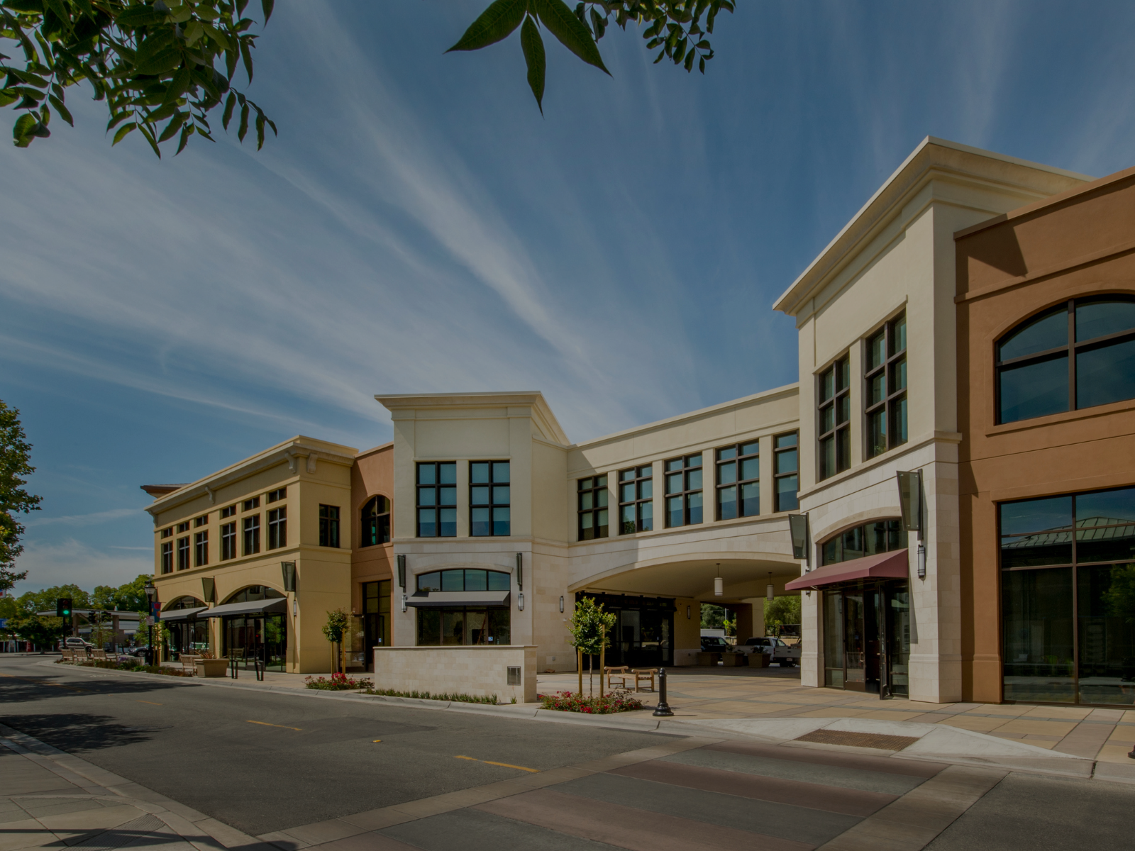A modern shopping plaza with storefronts, large windows, and an open street view on a sunny day with a blue sky and wispy clouds.