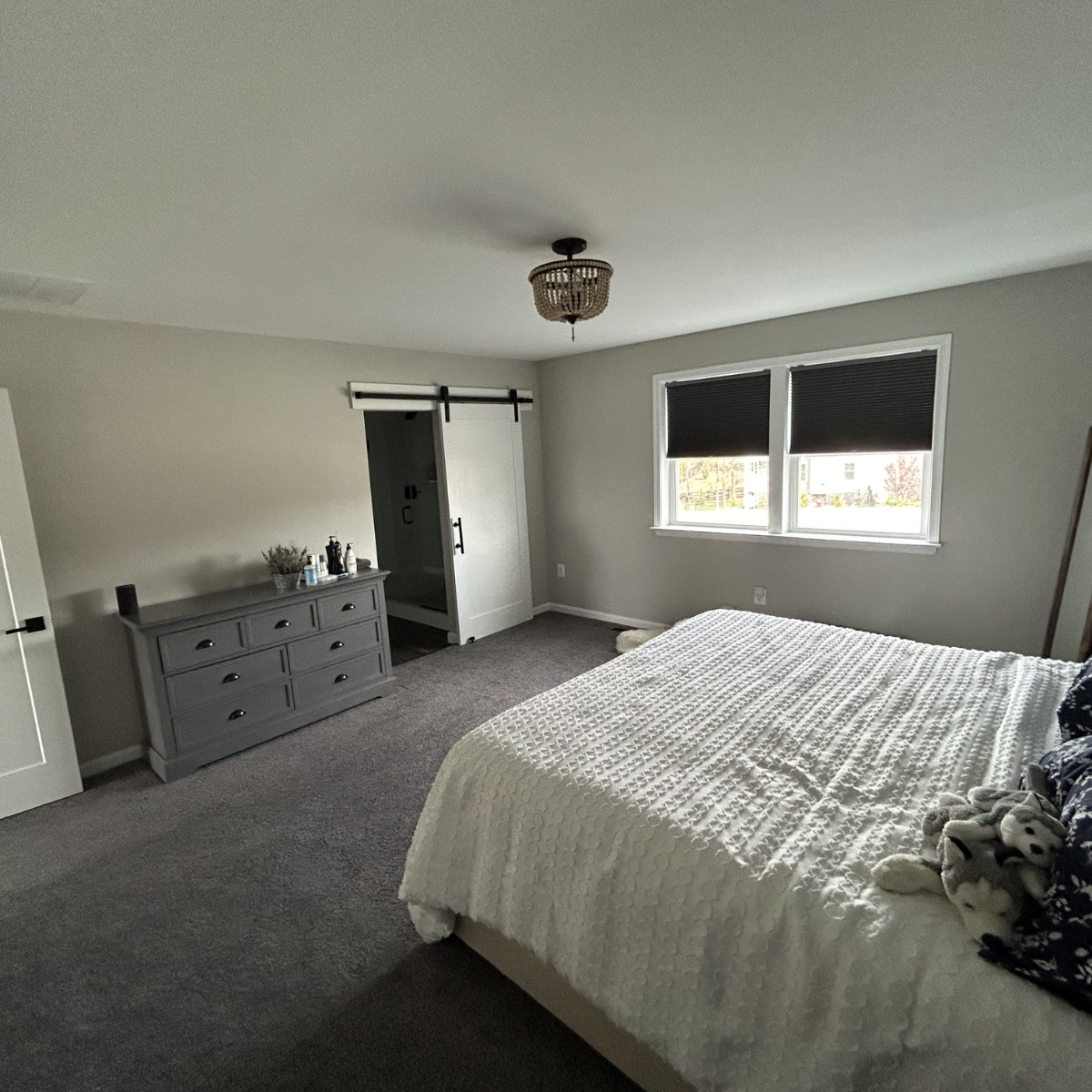A bedroom with a gray dresser, a bed with a white quilt, and a window with black blinds.