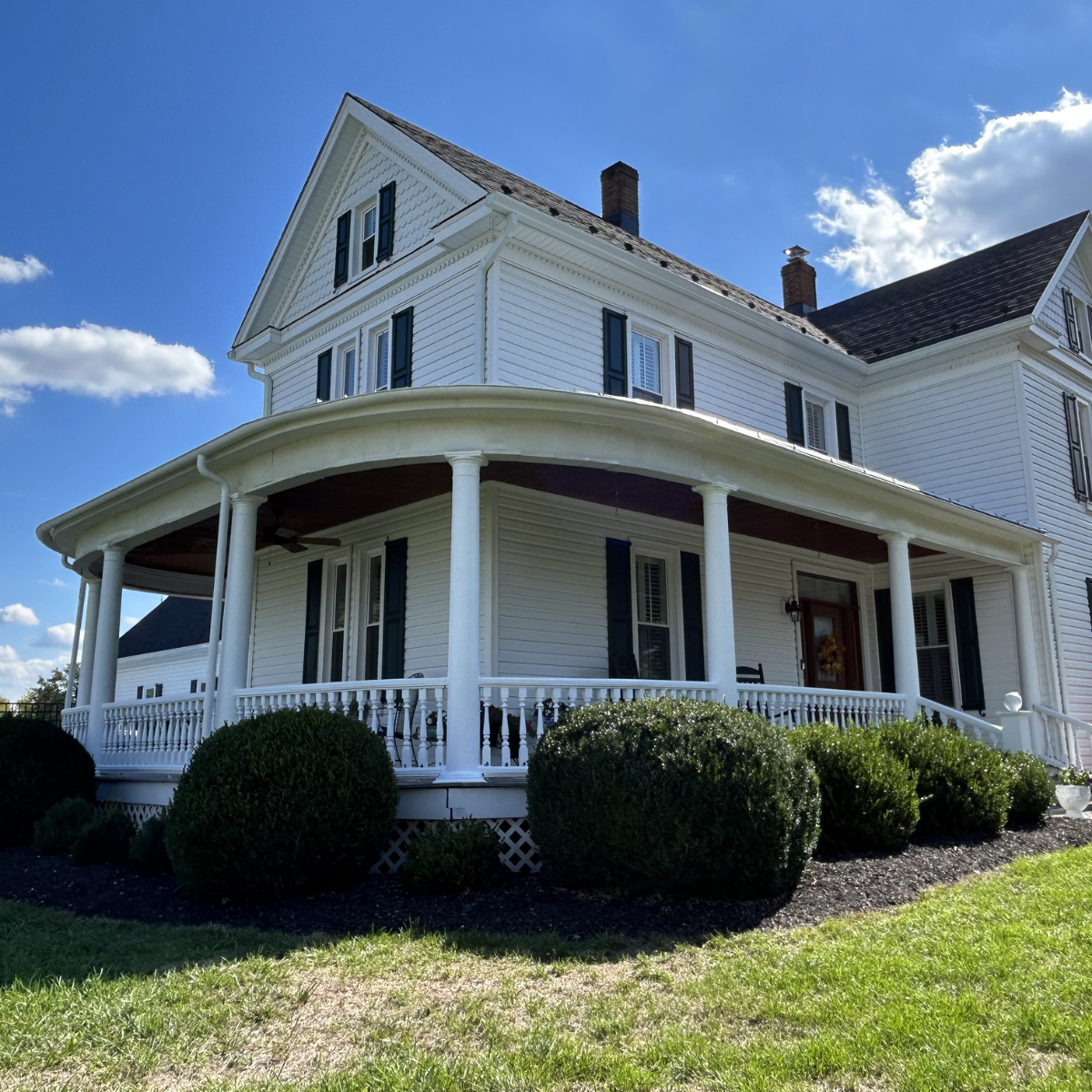 Expert exterior painters near you in Winchester, VA. Project: A large white Victorian-style house with a wraparound porch, black shutters, and a sloped roof, surrounded by manicured bushes and a green lawn under a partly cloudy blue sky.