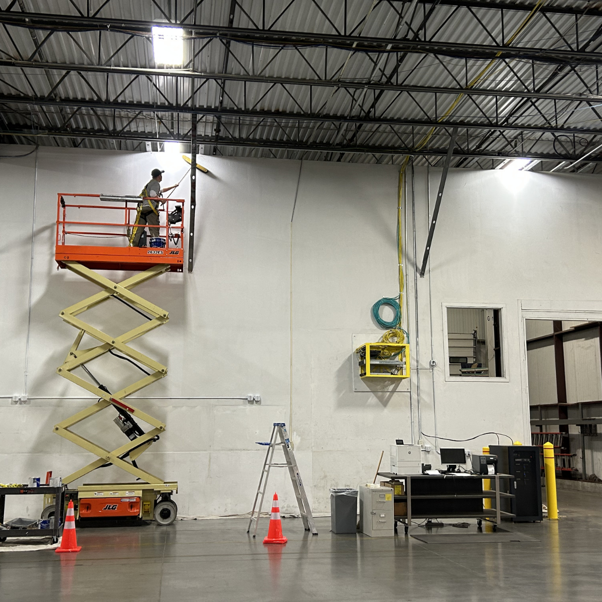 A worker on an orange scissor lift painting or cleaning a wall inside an industrial warehouse. The warehouse has a metal roof, fluorescent lighting, utility equipment, and tools on the floor. Commercial painting services Winchester Virginia 