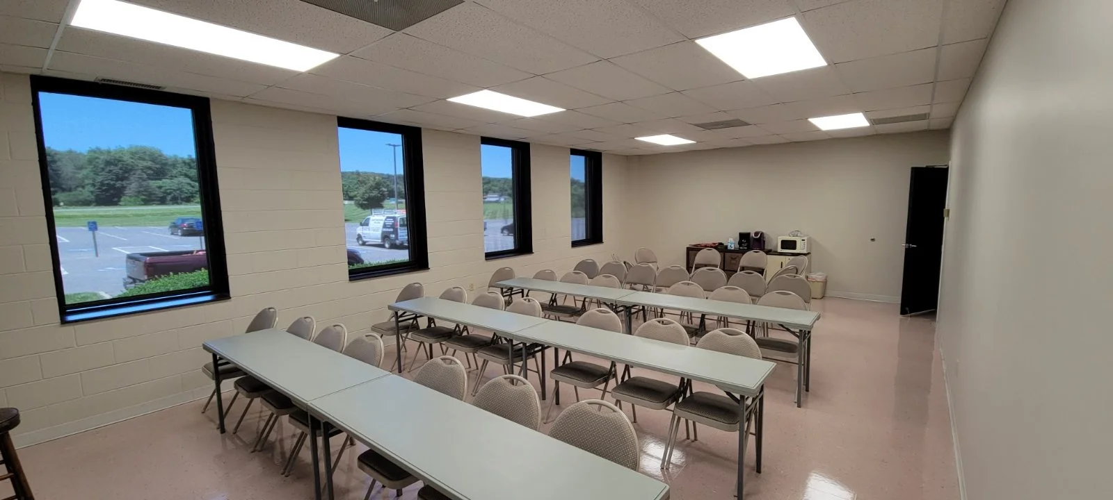 Commercial painters Conference or meeting room with three long white tables, beige chairs around them, and a kitchenette in the back with a microwave, coffee maker, and popcorn machine. Four windows on one wall show a parking lot and trees outside.