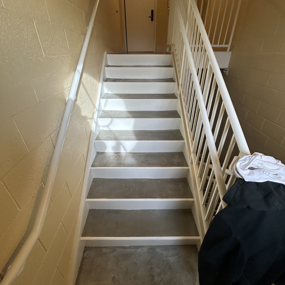 Indoor stairway with beige brick wall on the left, white railings on both sides, and gray steps with white risers. The top of the staircase leads to a closed door. Commercial painting services Winchester Virginia 