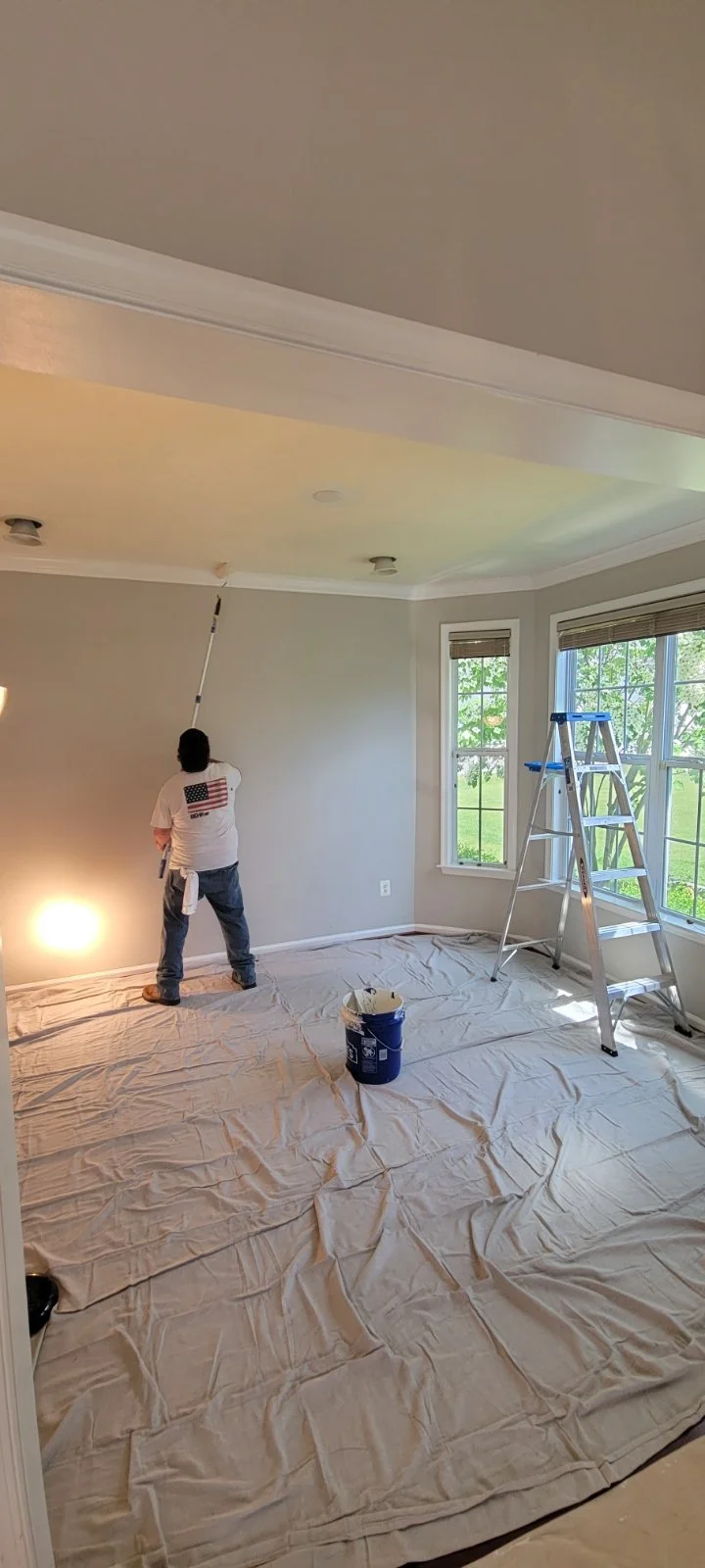 Person painting a wall with a roller in a room with large windows and a ladder, covered floor for painting prep. Interior painting Winchester Virginia