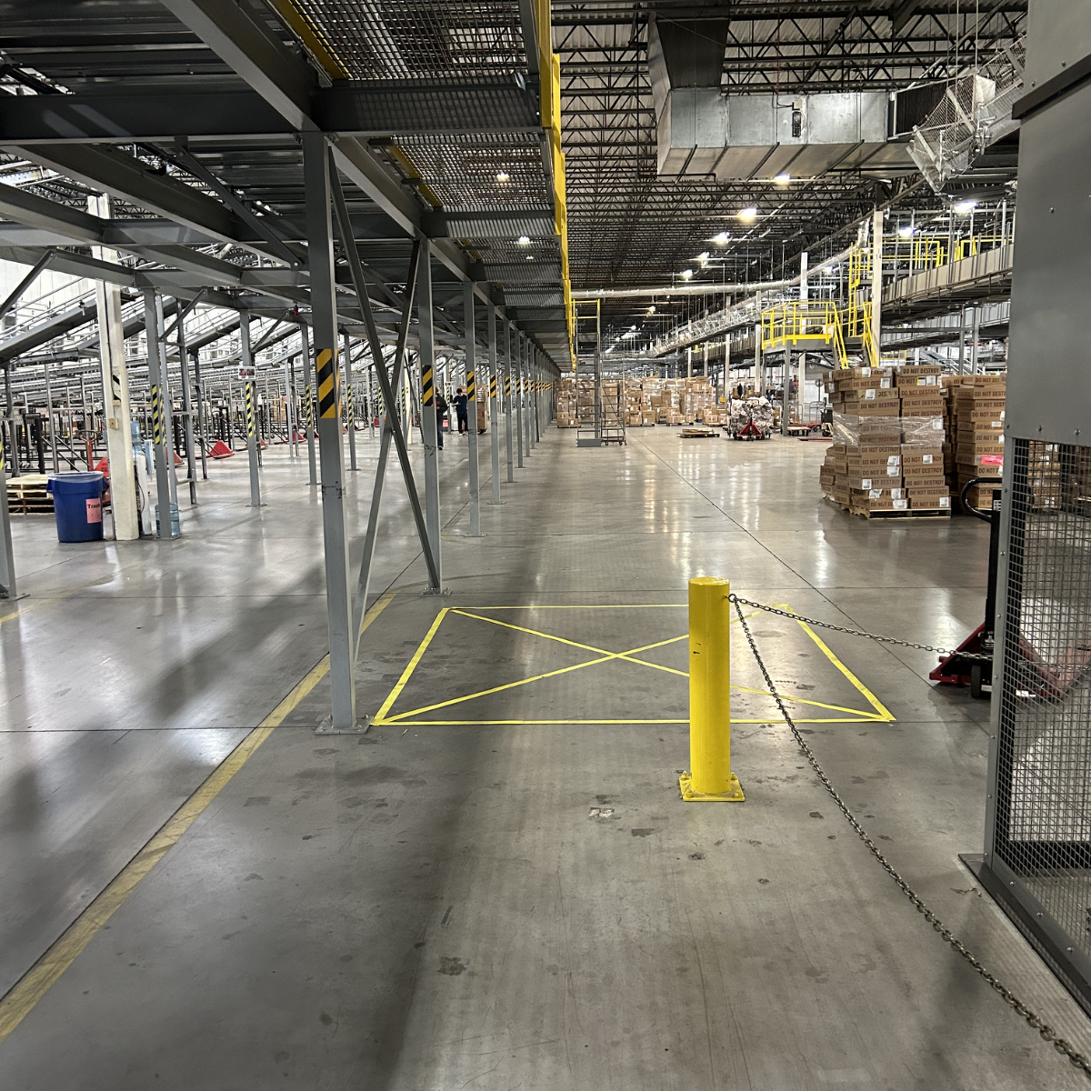 An empty warehouse or distribution center with metal shelving, pallets of boxes, and worker employees in the background. There are yellow safety barriers and a chain link fence in the foreground.