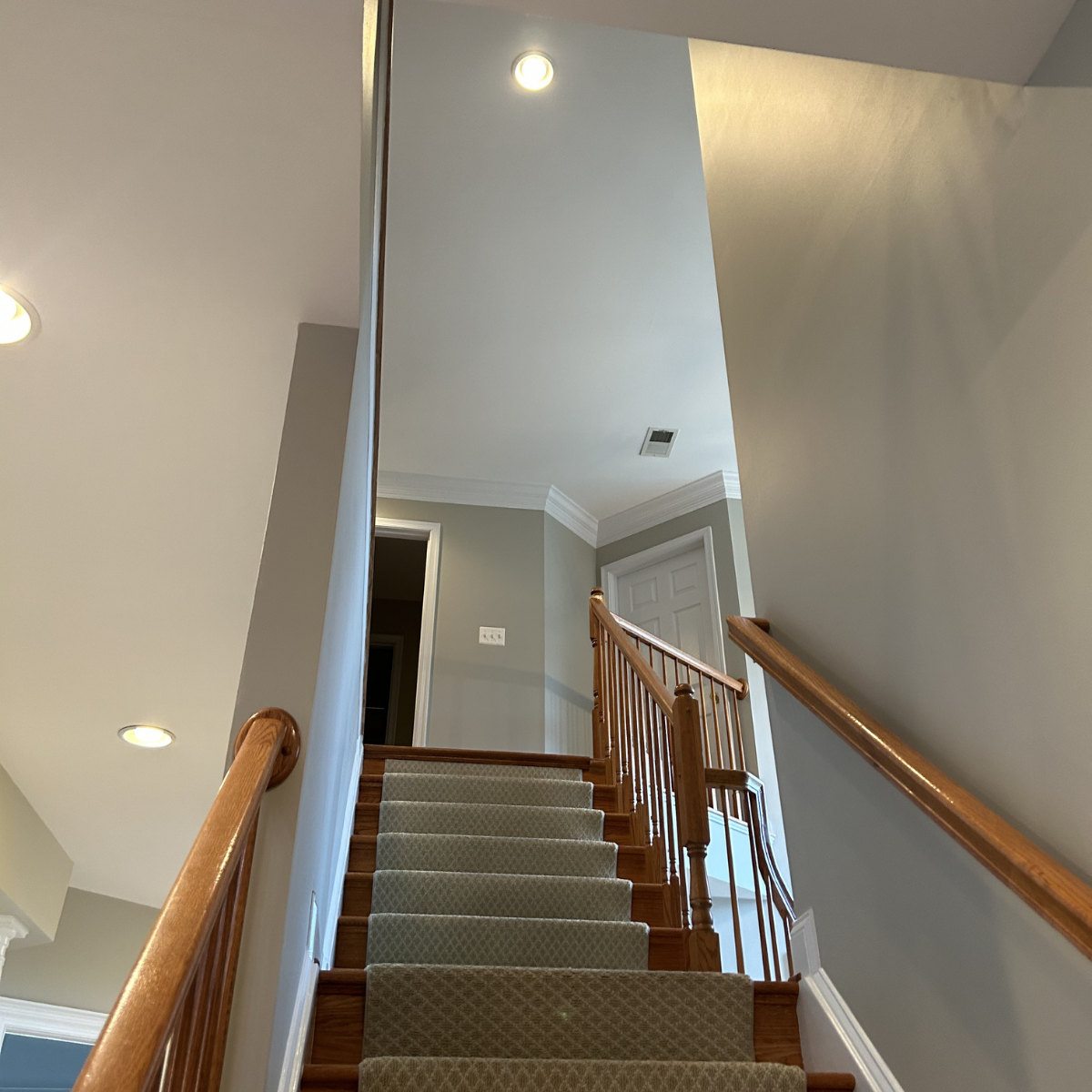Photo of a staircase with wooden handrails, carpeted steps, leading to a hallway with closed white door, beige walls, and ceiling lights.