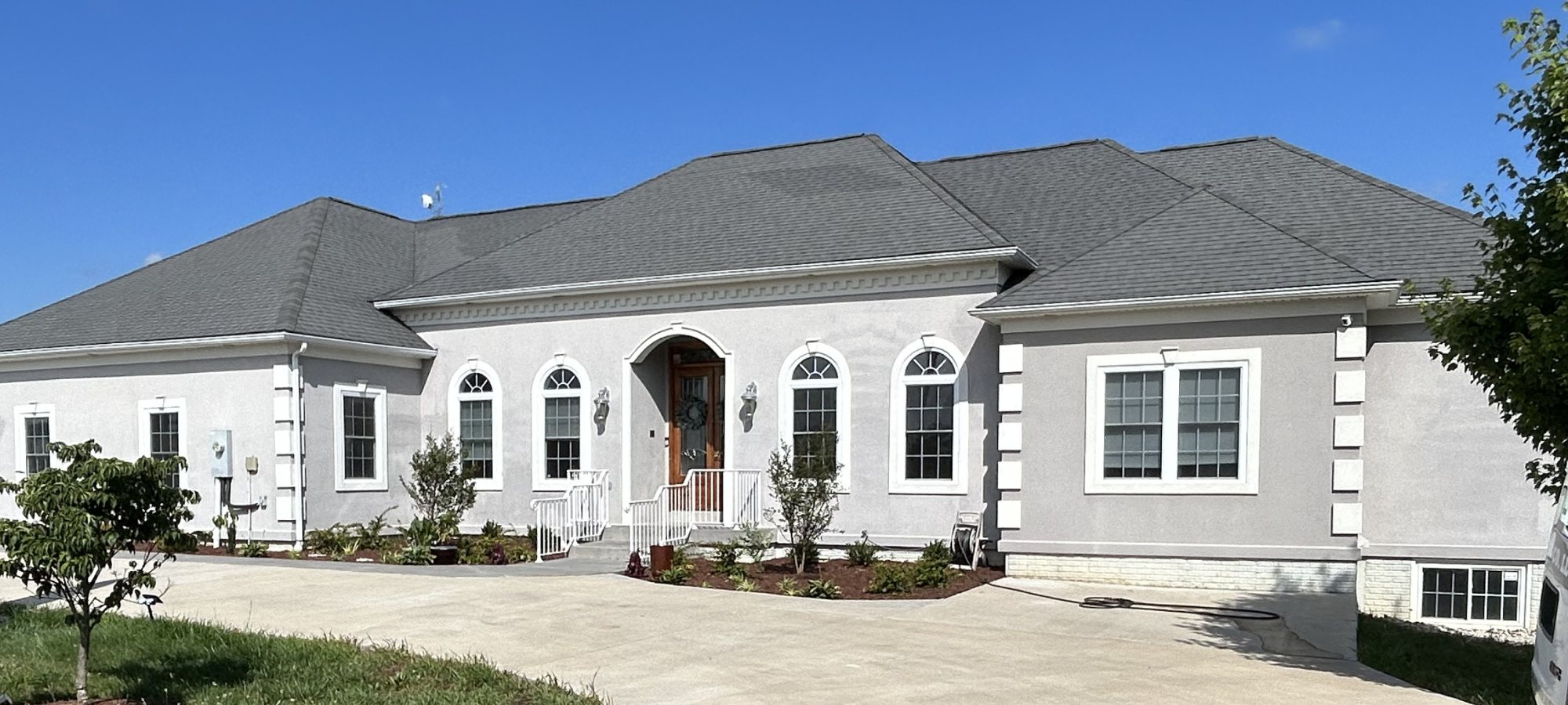 CMT Painting Co. exterior house painters Winchester VA – A modern, white, single-story house with an arched wooden door, surrounded by a driveway and landscaped front yard with small trees and plants, under a clear blue sky.