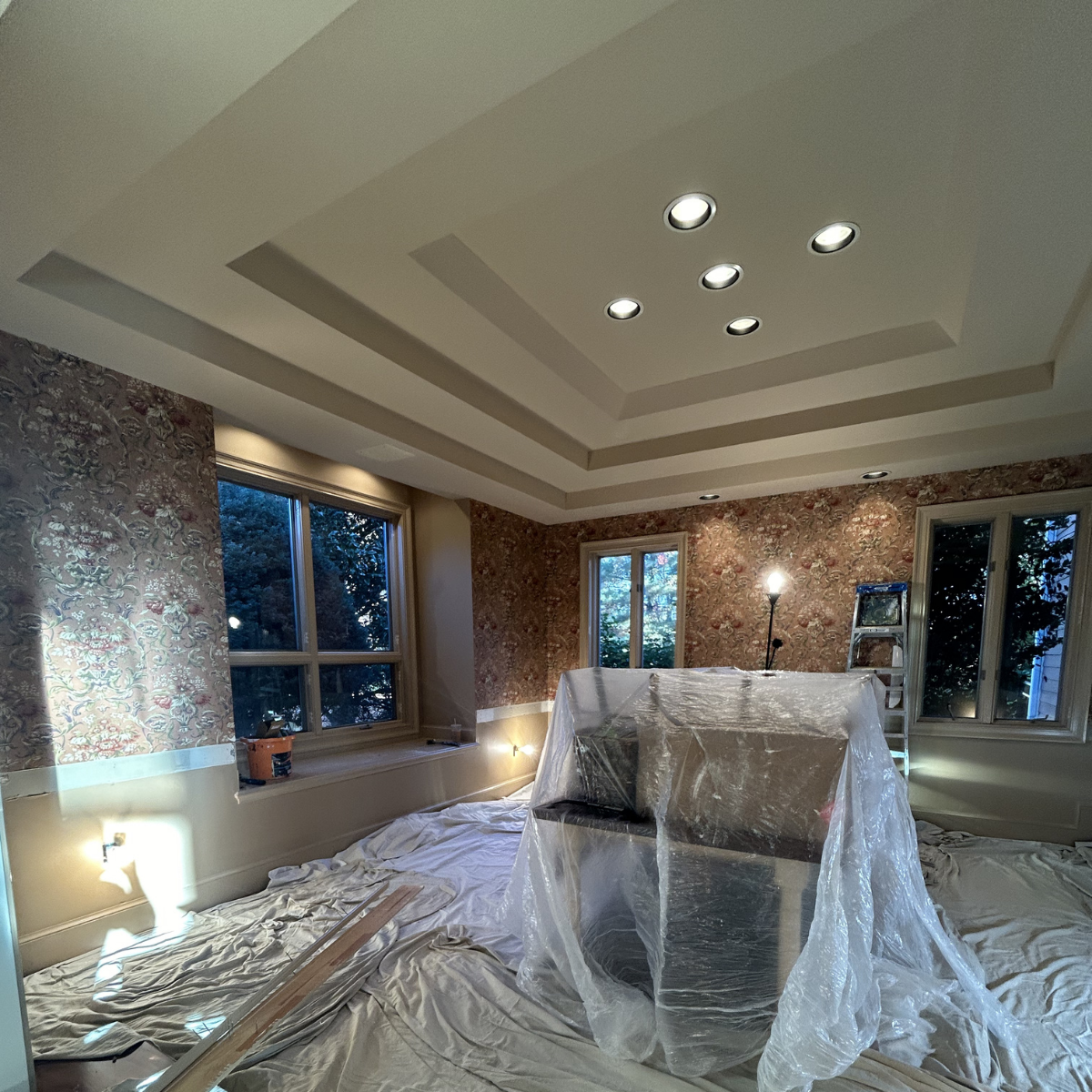 Living room under renovation with plastic-covered furniture, construction lighting, and decorative wallpaper on the walls.