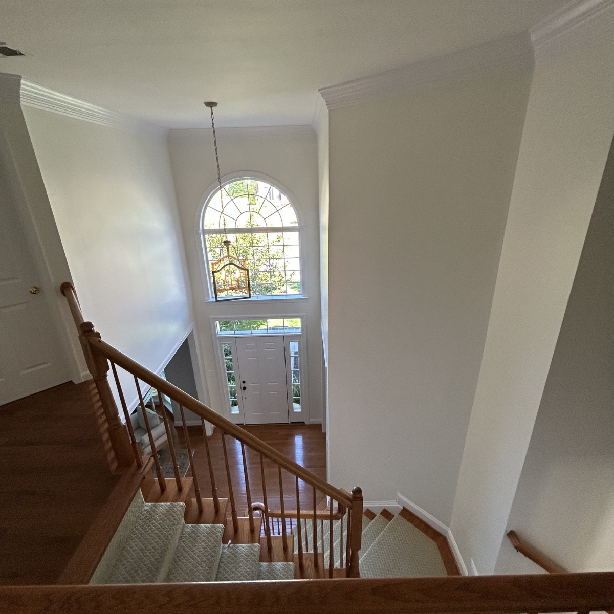 Interior view of a house entrance with a staircase, front door, large arched window, and foyer with hardwood floors.