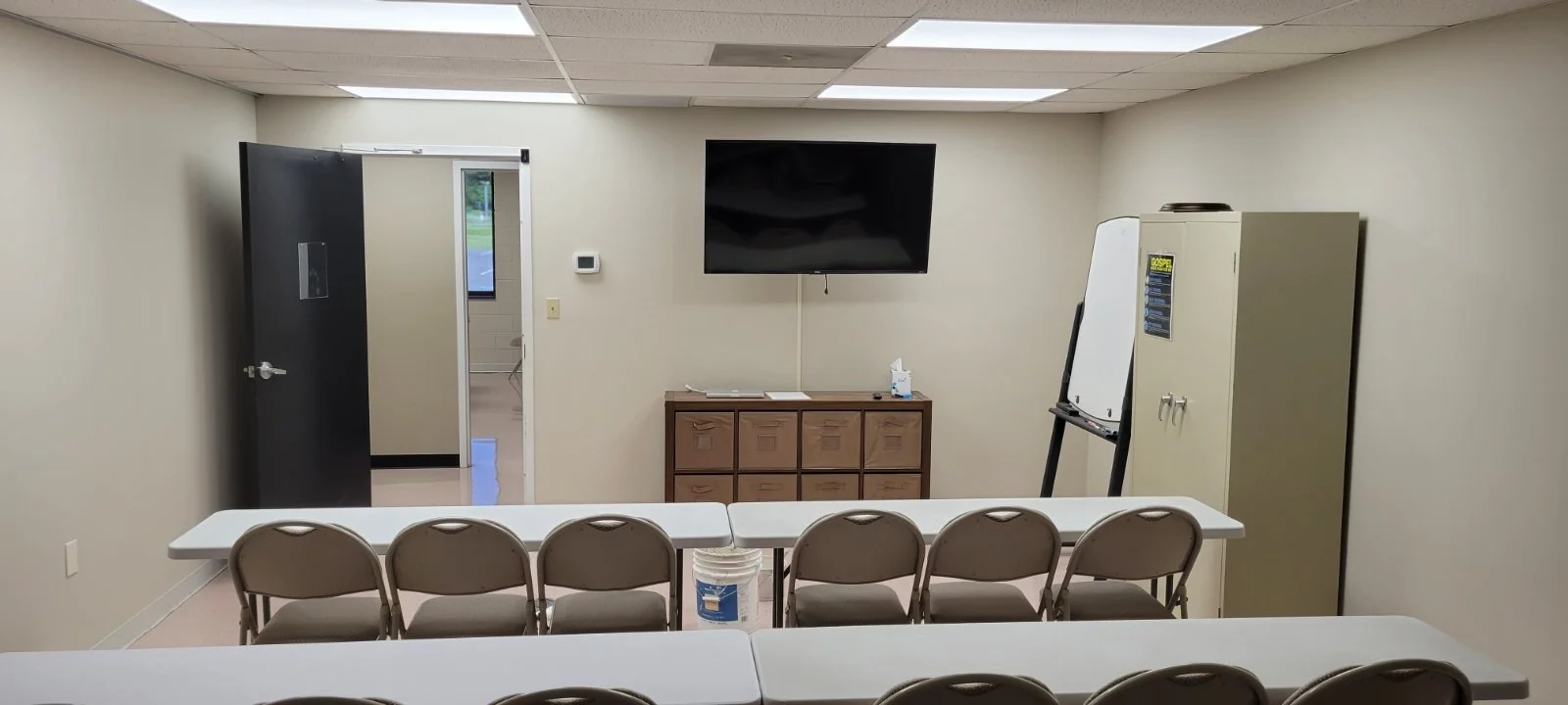 Empty classroom or meeting room with rows of chairs, a black TV on the wall, white tables, a wooden cabinet, a whiteboard, a closet, and an open door leading to another room.