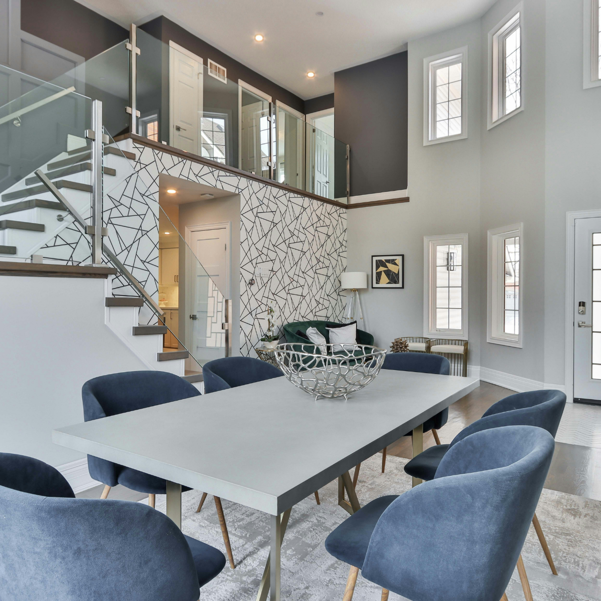 Modern dining room with a white table, eight blue velvet chairs, a decorative silver bowl, and a gray rug. The walls feature multiple rectangular windows, and a staircase with glass and metal railing leads to an upper floor. A geometric black and white accent wall is visible near the staircase.