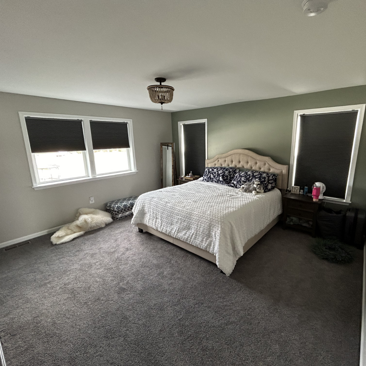 A bedroom with a large bed with a upholstered headboard, dark patterned pillows, and a white textured blanket. There are three windows with black blinds, a nightstand with items including a pink water bottle and a blanket, and a full-length mirror. A