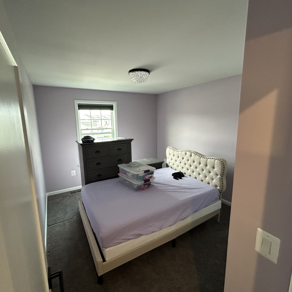 Bedroom with purple walls, a bed with a tufted headboard, dresser, and window, with some clothes and boxes on the bed.