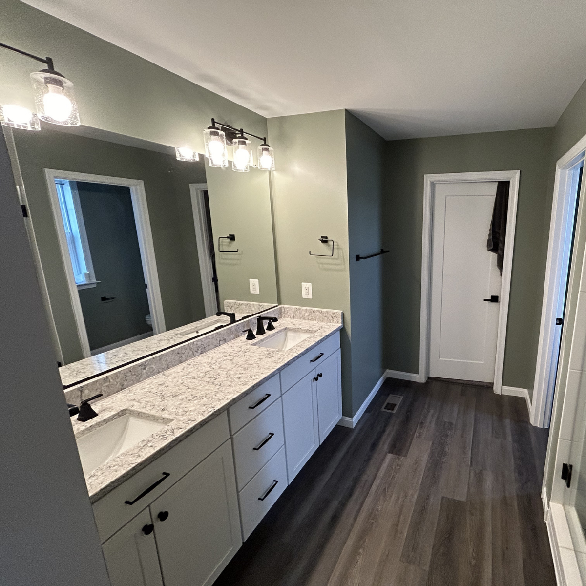 Bathroom with double vanity, granite countertop, black fixtures, large mirror, green walls, dark wood flooring, and four light fixtures above mirror.