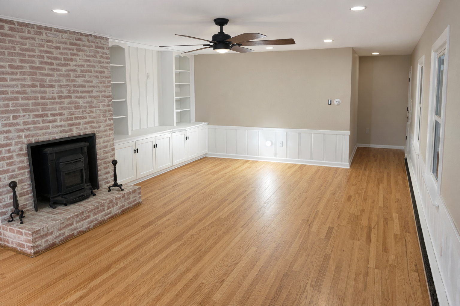 After a complete repaint of a living room in Winchester VA by CMT Painting Co. Empty living room with hardwood floor, brick fireplace, white built-in cabinets, beige walls, and ceiling fan