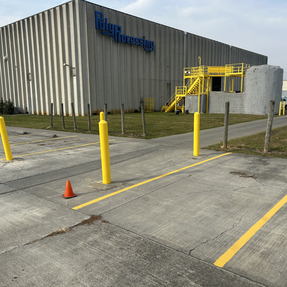 Commercial painting Industrial warehouse building with a sign that says "PolyProcessing," yellow safety bollards, and a yellow staircase leading to a platform on the side of the building. Parking lot with yellow lines and a small orange traffic cone.