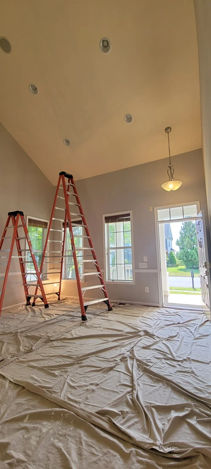 Interior painting Winchester Virginia Room with ceiling lights, a hanging light fixture, windows, a door, ladders, and a plastic sheet covering the floor.