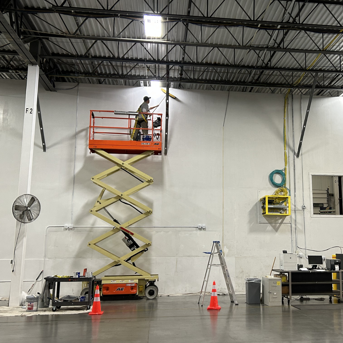 Commercial Painting Winchester, VA by CMT Painting Co. A worker standing on an orange scissor lift painting a ceiling light fixture in an industrial or commercial space.