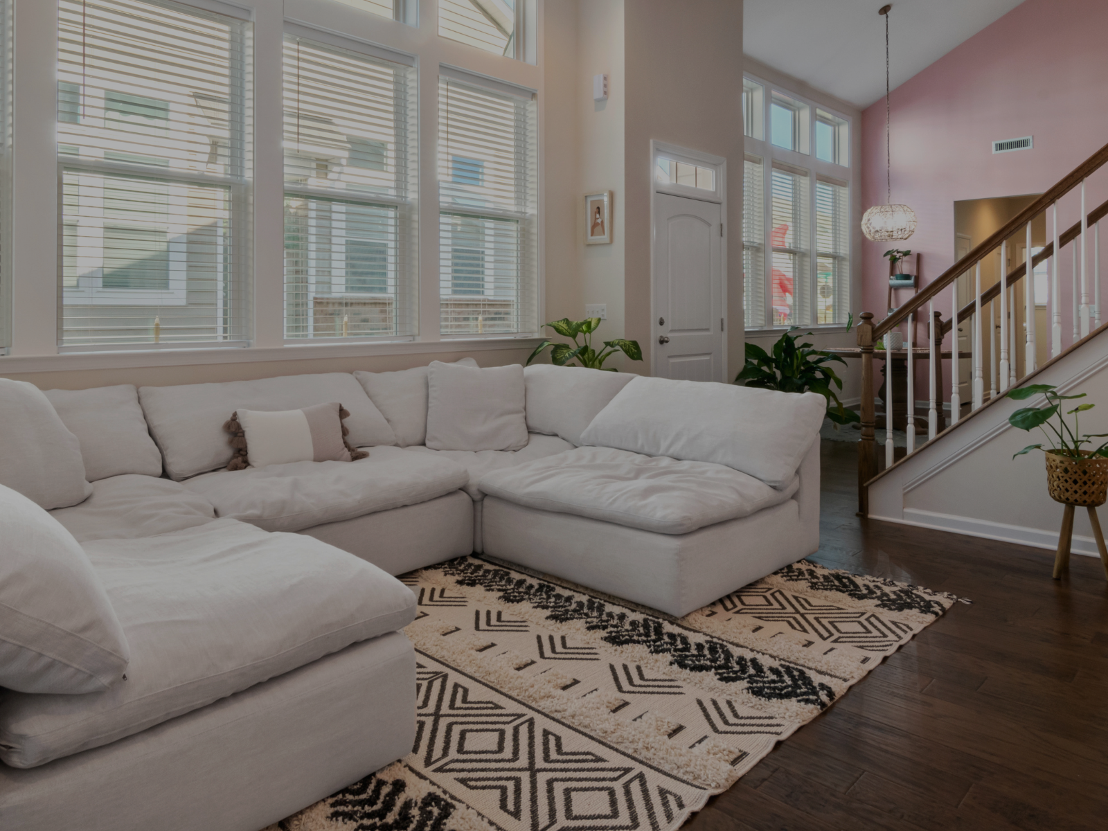 Living room with a white sectional sofa, patterned rug, large windows with blinds, and a staircase with wooden railings.