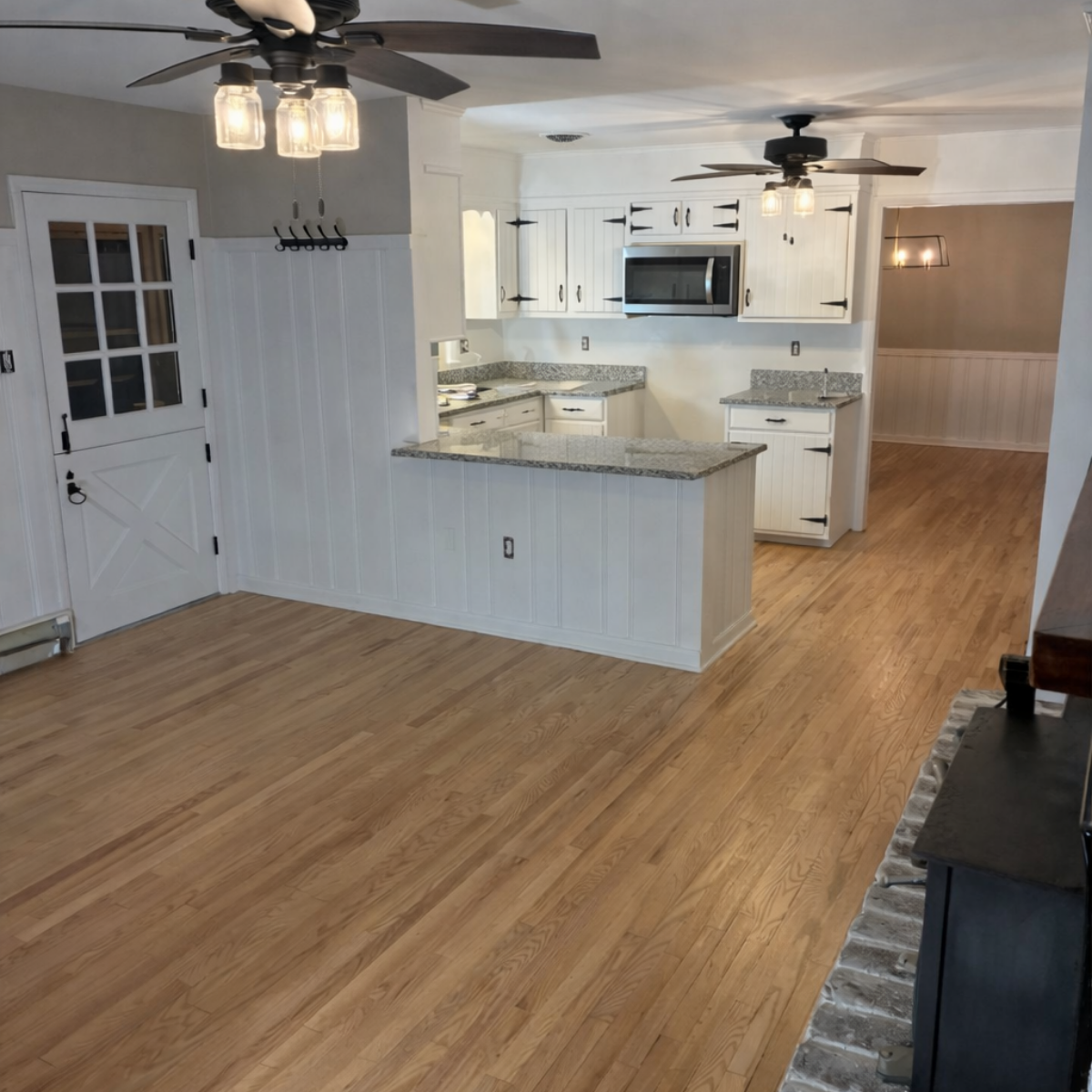 Residential Painting by CMT Painting Co. Empty living area with hardwood floors and an open kitchen with white cabinets, granite countertops, stainless steel microwave, and ceiling fans with lights.