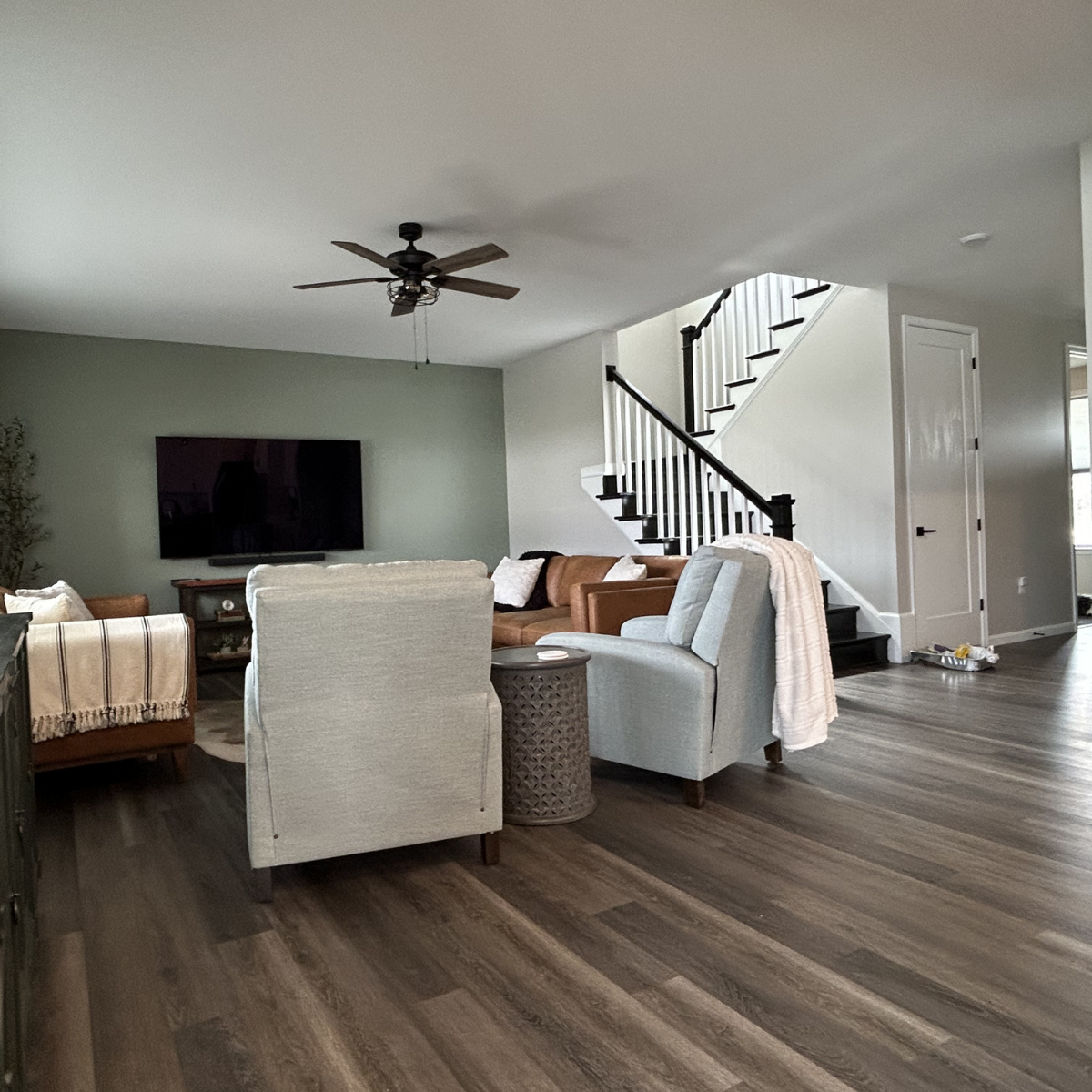 Living room with a mounted TV, a ceiling fan, beige and brown furniture, and a staircase leading upstairs. The room has wood flooring and neutral-colored walls.