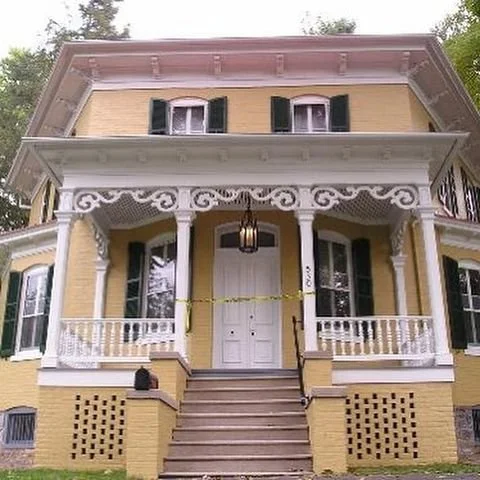 Yellow two-story house with white trim and black shutters, front porch with decorative white railing, steps leading to white front door, and ornate white woodwork under the porch roof.
