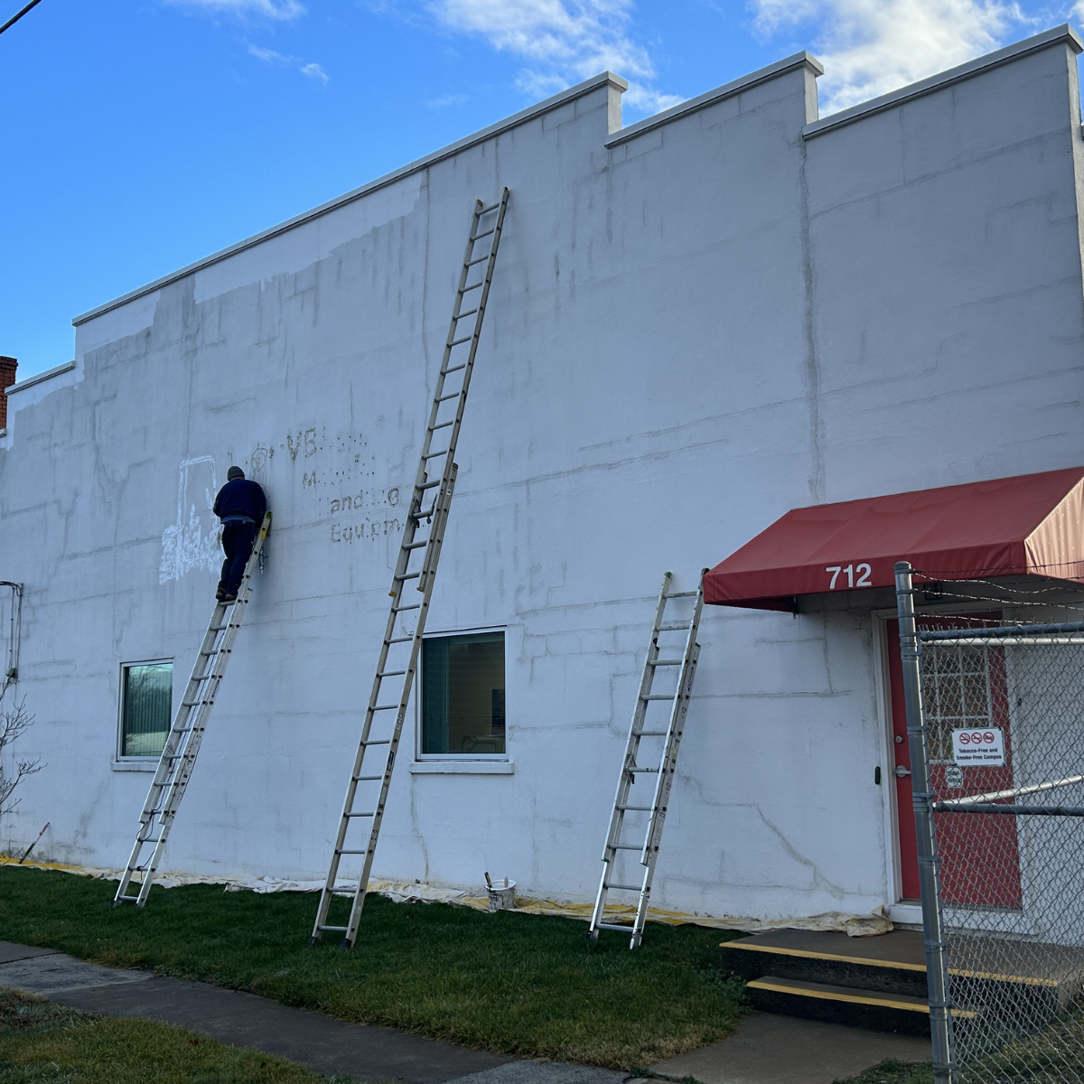 A worker using a ladder to paint or write on the white exterior wall of a building, with two additional ladders leaning against the wall. The building has two small windows and a red awning over the door labeled 712. The sky is blue with some clouds.