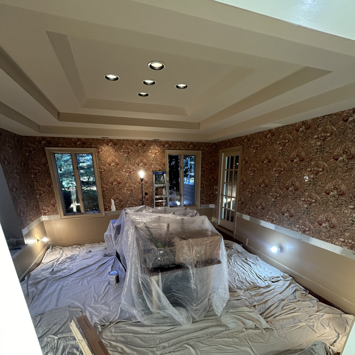 Living room under renovation with plastic-covered furniture, a step ladder, and construction lights, featuring floral wallpaper and a tray ceiling with recessed lighting.