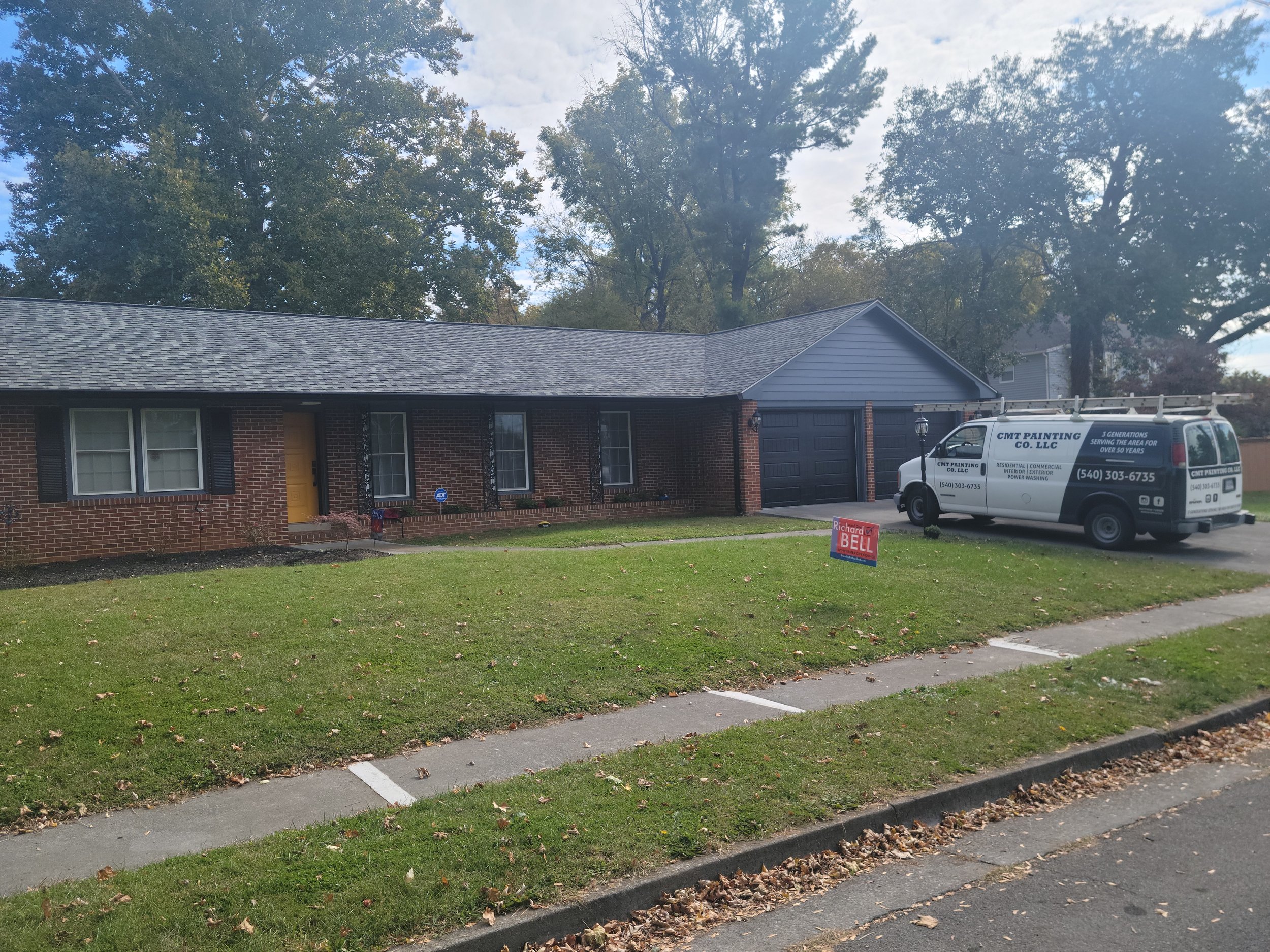 CMT Painting Co. exterior house painters Winchester VA – Brick house with yellow door and black shutters, front yard with a small sign, driveway with all-purpose van, and trees in the background.