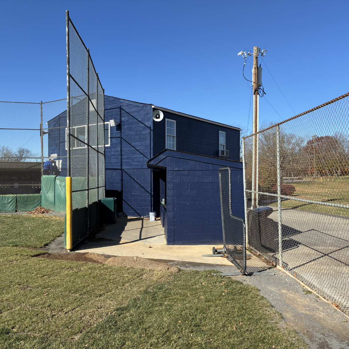CMT Painting Co. commercial painting Northern Virginia – Blue building with small door, surrounded by chain-link fences, with baseball field and person in the background, on a clear sunny day.