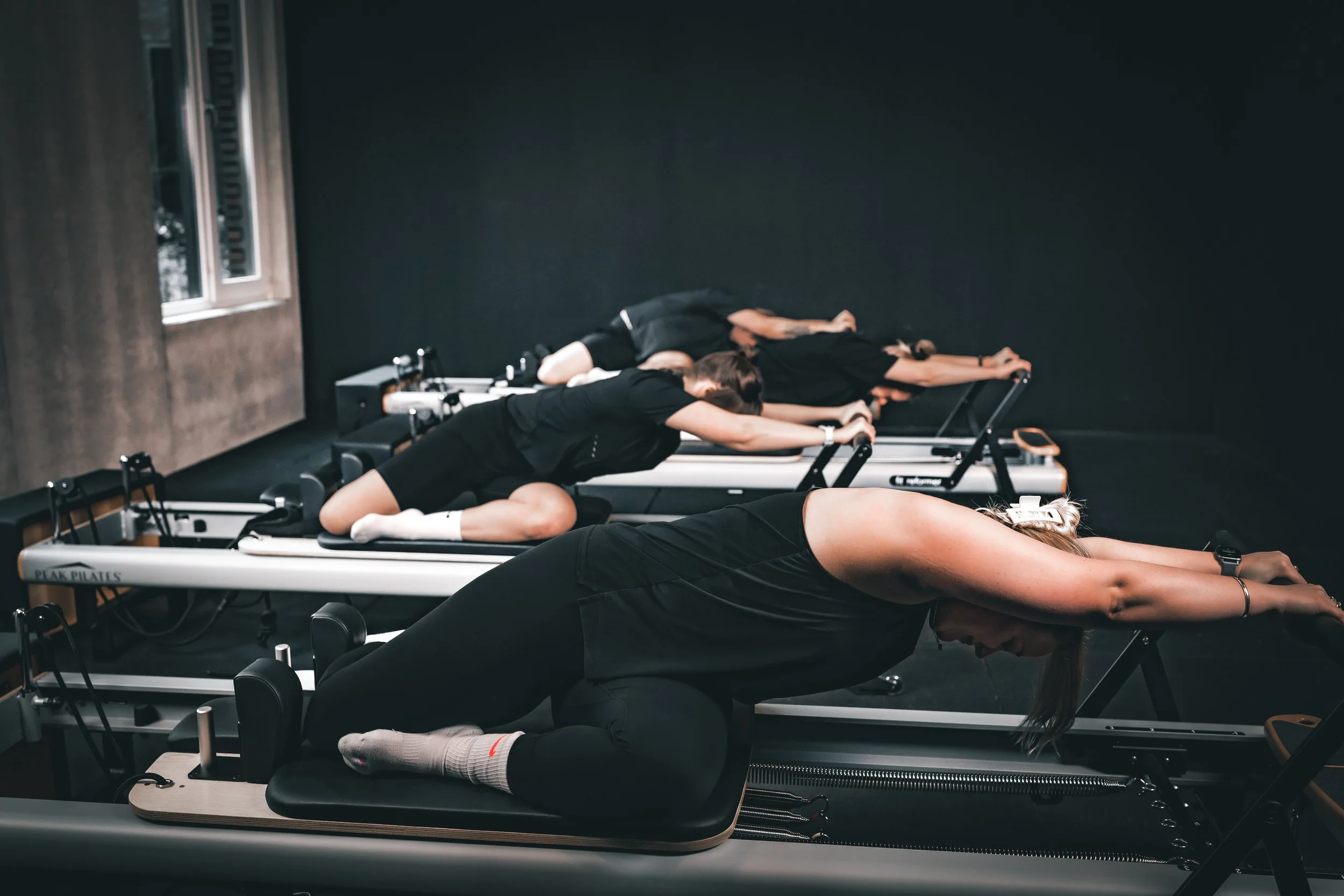 Four women doing Pilates on reformer machines in a dark studio.