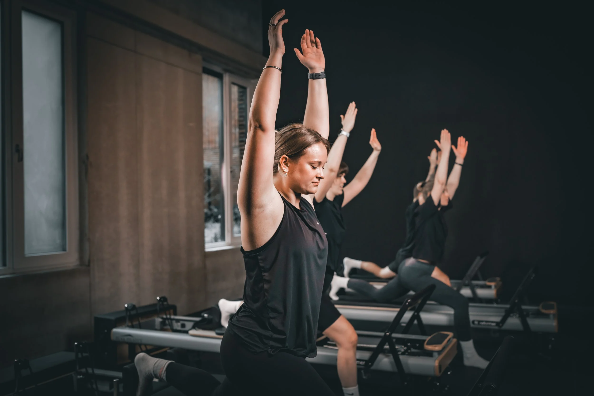 Three women in exercise attire participate in a workout class using reformer machines in a dark gym room with large windows.