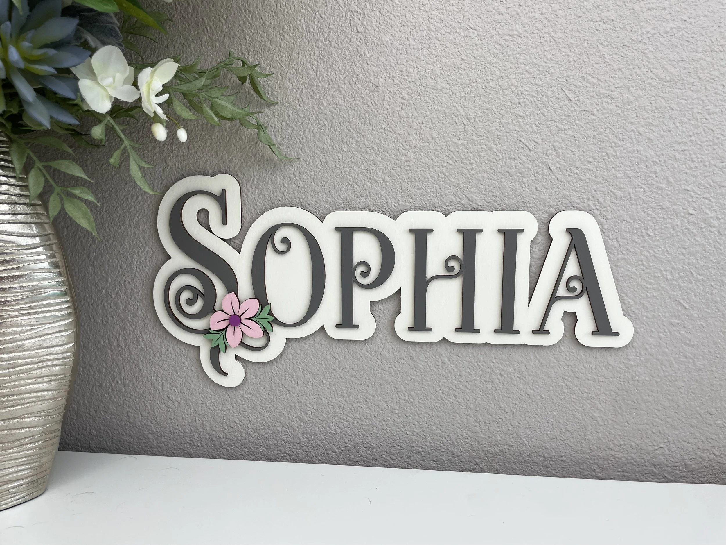 Decorative sign that says 'SOPHIA' with a pink flower and green leaves attached to the letter 'S,' placed on a textured light gray wall near a silver vase with white flowers and greenery.