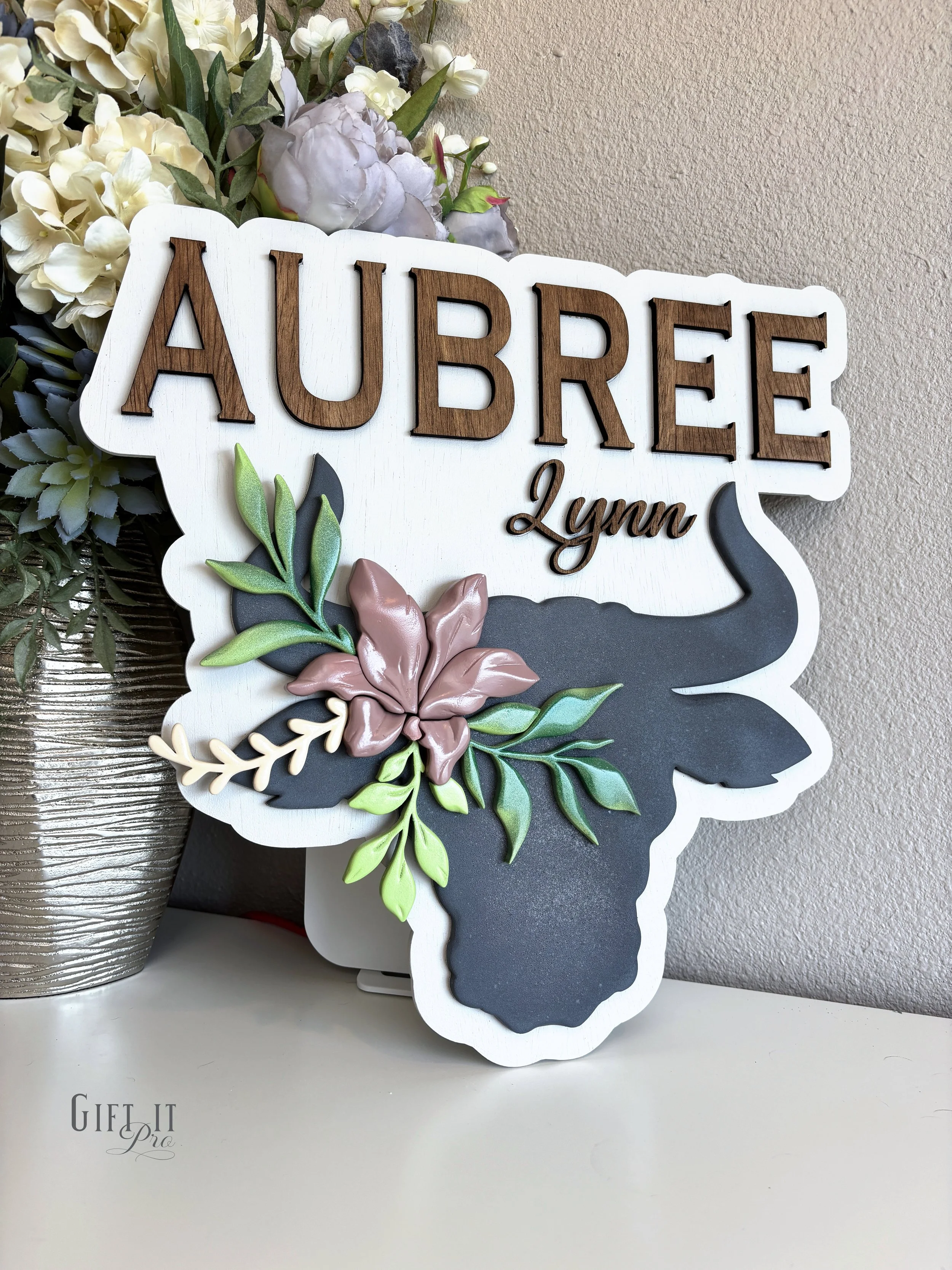 Decorative sign with the word 'AUBREE' in large wooden letters, 'Lynn' in smaller script, and a silhouette of a longhorn with floral accents, placed next to a bouquet of flowers in a silver vase.
