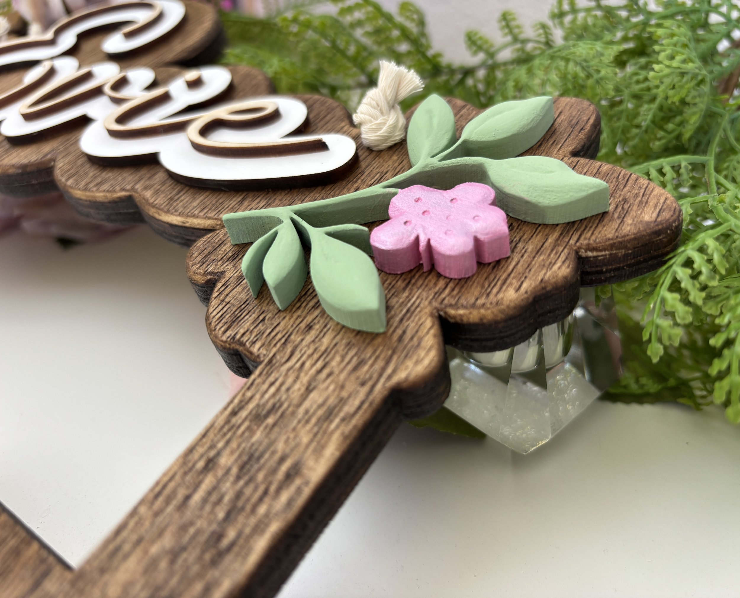Close-up of a wooden sign with the word 'Love' on it, decorated with pastel-colored wooden flowers and green leaves, surrounded by green foliage.