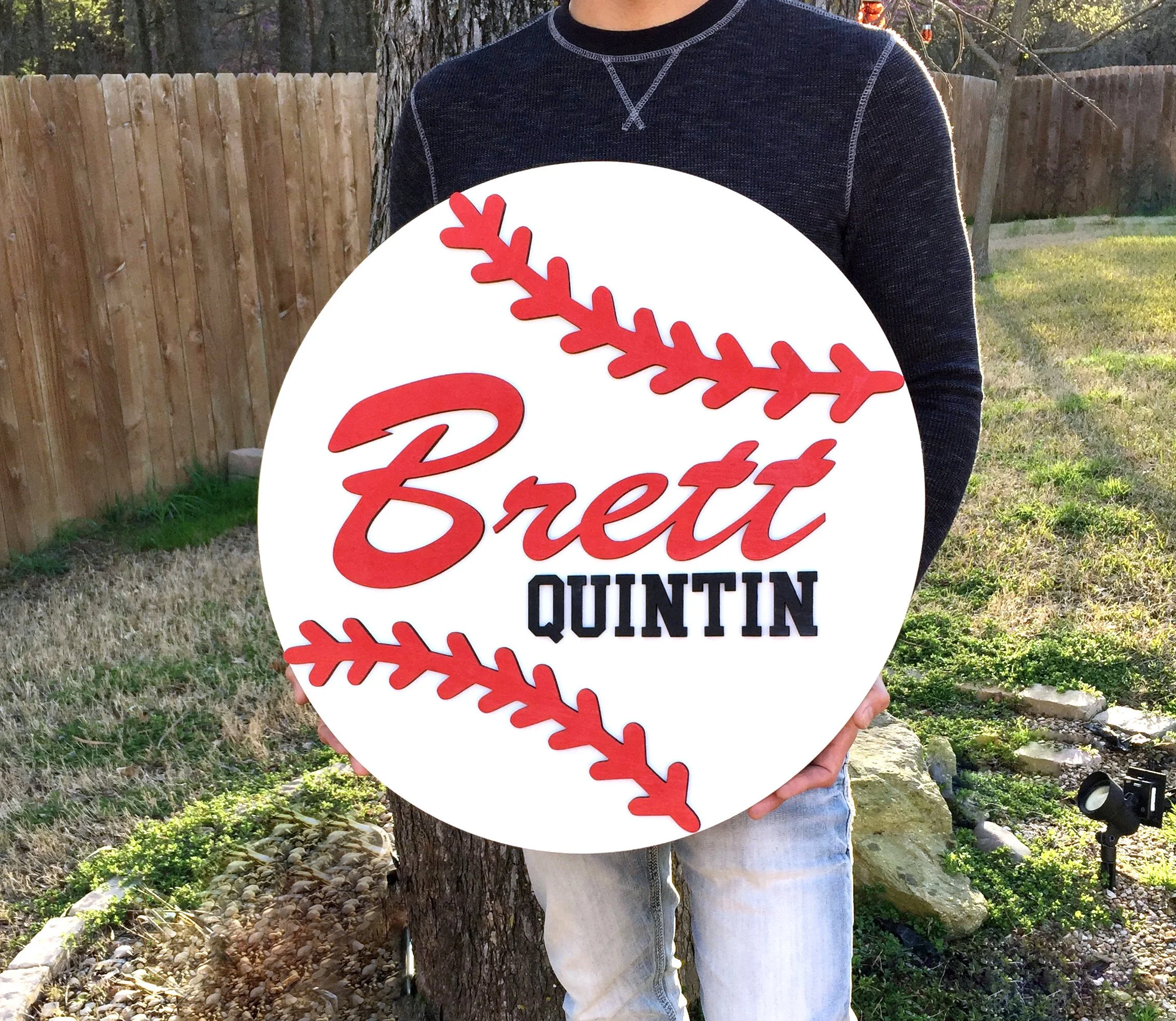 Person holding circular sign with a red baseball seam design, displaying the name Brett Quintin in red and black lettering, outdoors in a backyard with a wooden fence and grass.