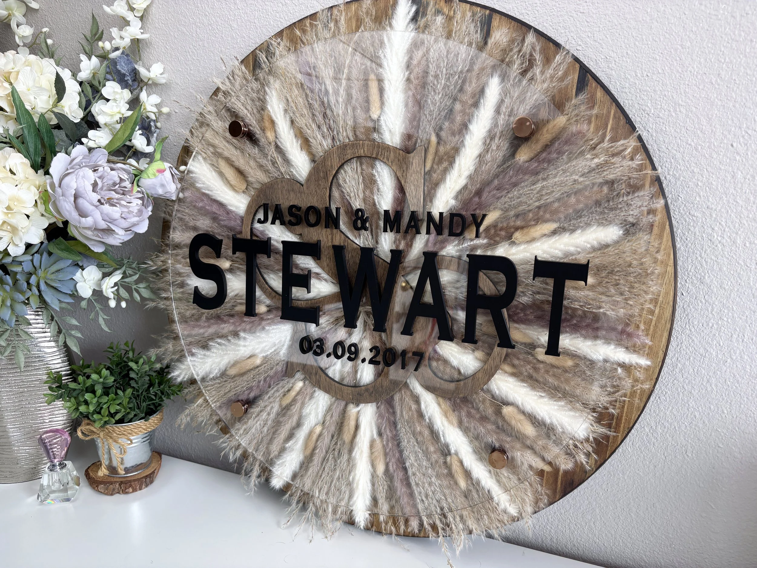 A decorative wooden and acrylic wall art piece with pampas grass and floral accents, engraved with the names Jason and Mandy, the date 03.09.2017, and the last name Stewart.