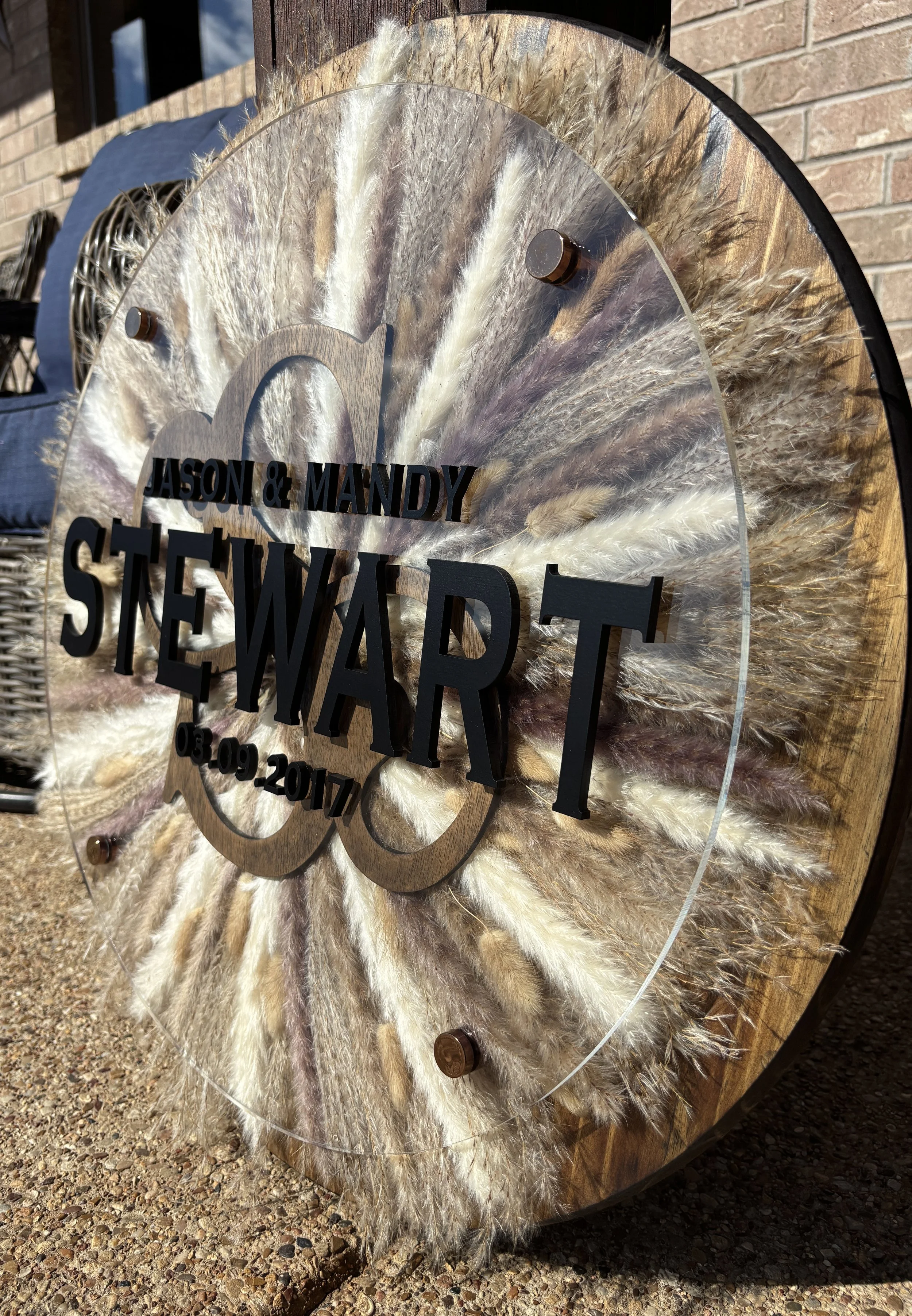 A decorative wedding sign with black raised lettering reading 'Jason & Mandy Stewart 03-09-2017,' mounted on a round wooden background with a clear acrylic front and feathered accents behind the lettering.