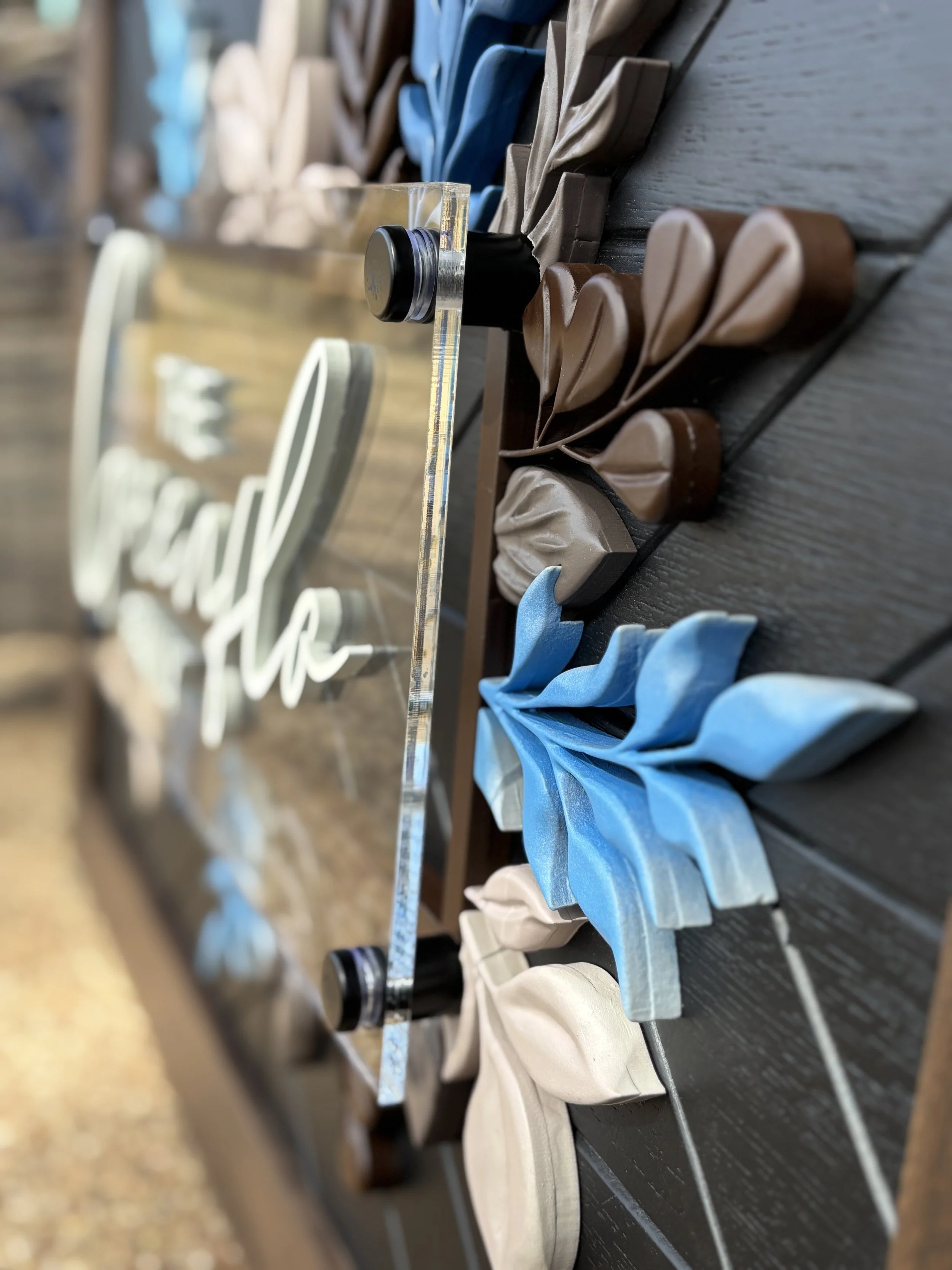 Decorative arrangement with hand carved flowers in shades of beige, brown, blue, and gray, placed on a dark wooden surface beside a clear acrylic sign with white cursive text.
