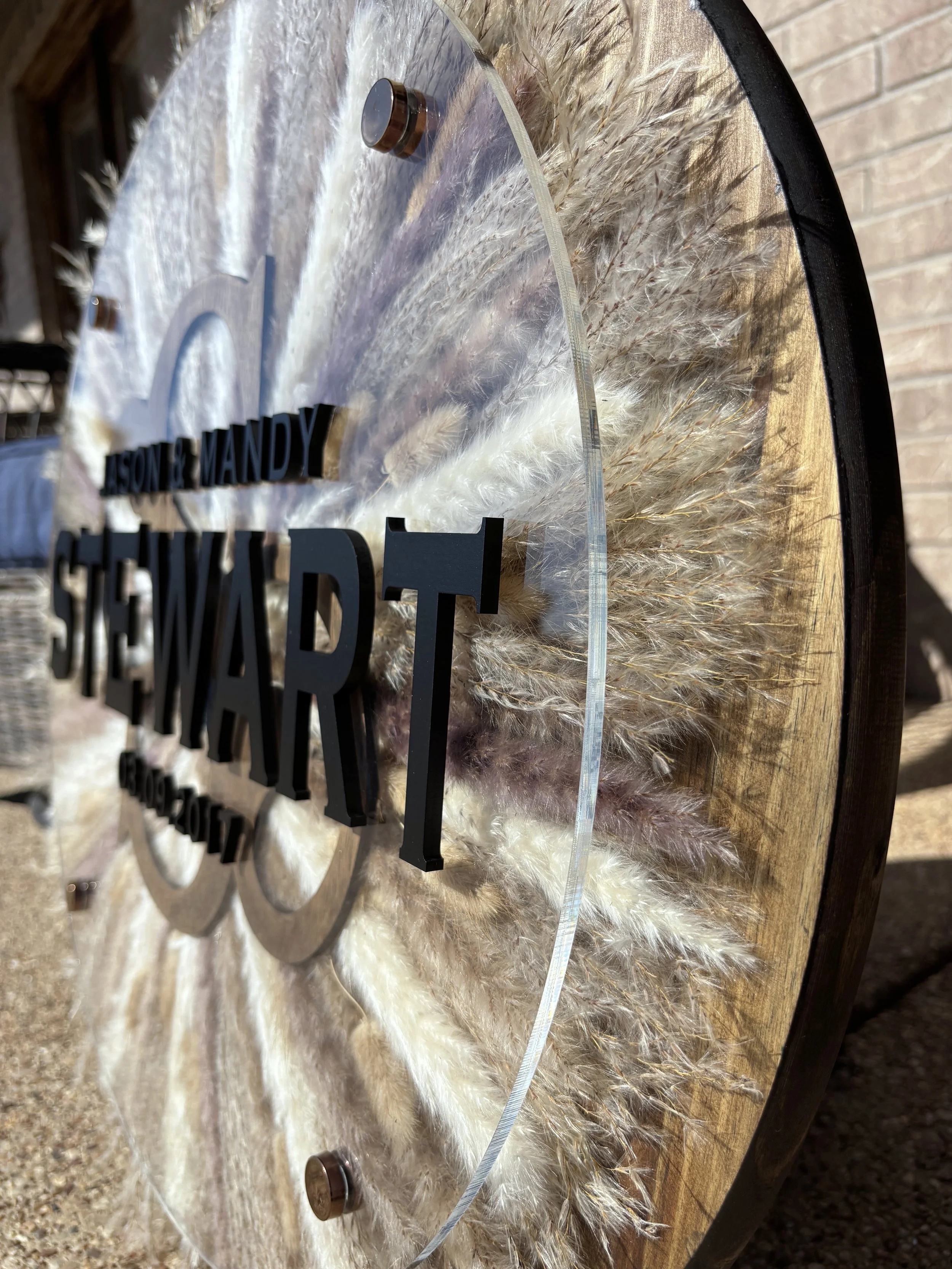Close-up of a decorative wall clock with black letters spelling out "STEwart" and other words, mounted on a round wooden and acrylic frame, outdoors on a sunny day.