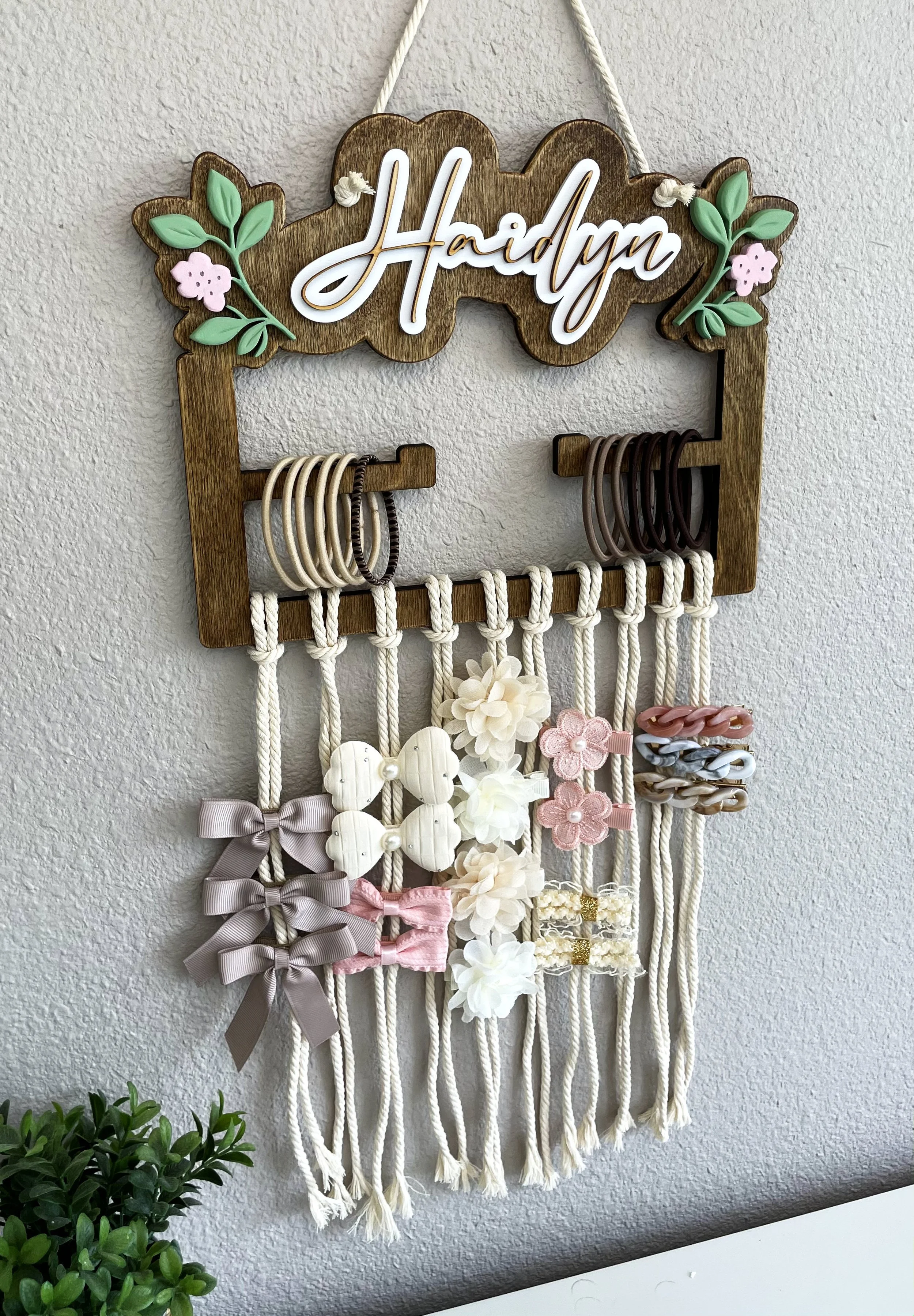 Wooden jewelry organizer with the name 'Haidyma' on a decorative sign at the top, displaying various hair accessories such as bows, scrunchies, hair clips, and headbands.