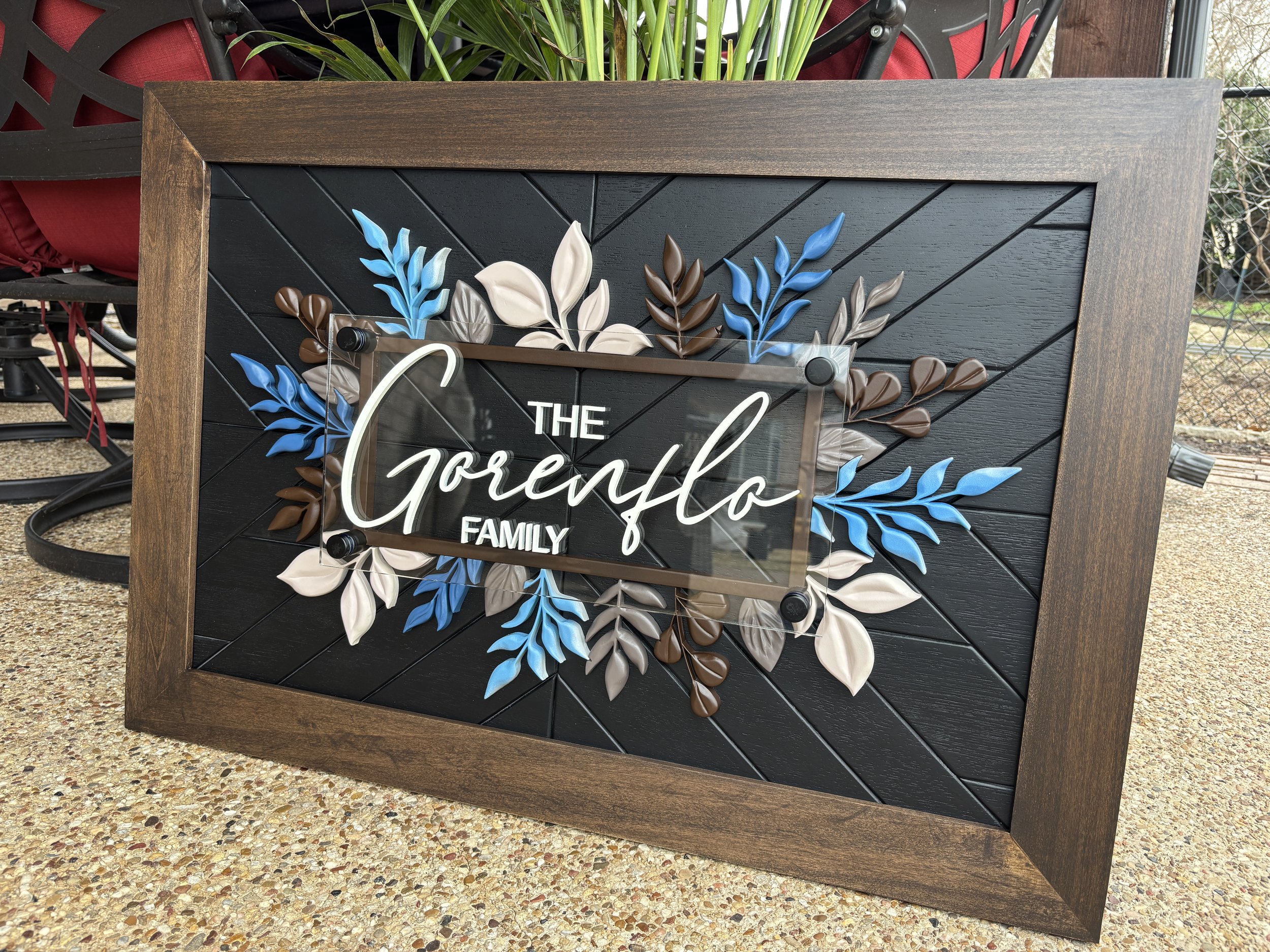 Framed decorative sign with the words ' Family last name'  and a floral design with blue, beige, and brown leaves, placed on a pebbled surface outdoors.